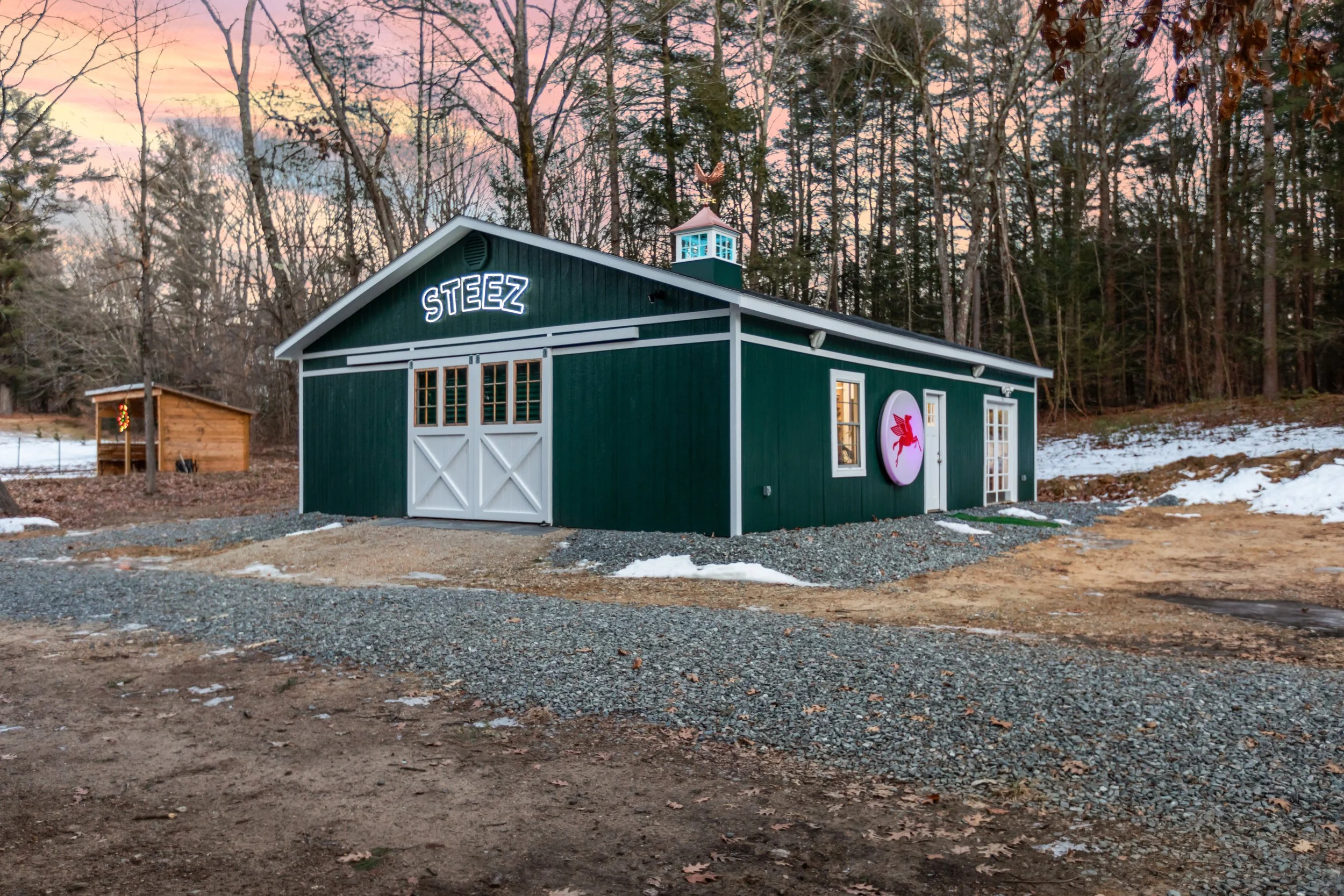Dark green building labeled “STEEZ” in a snowy woodland at sunset, with white doors, cupola, and illuminated sign.