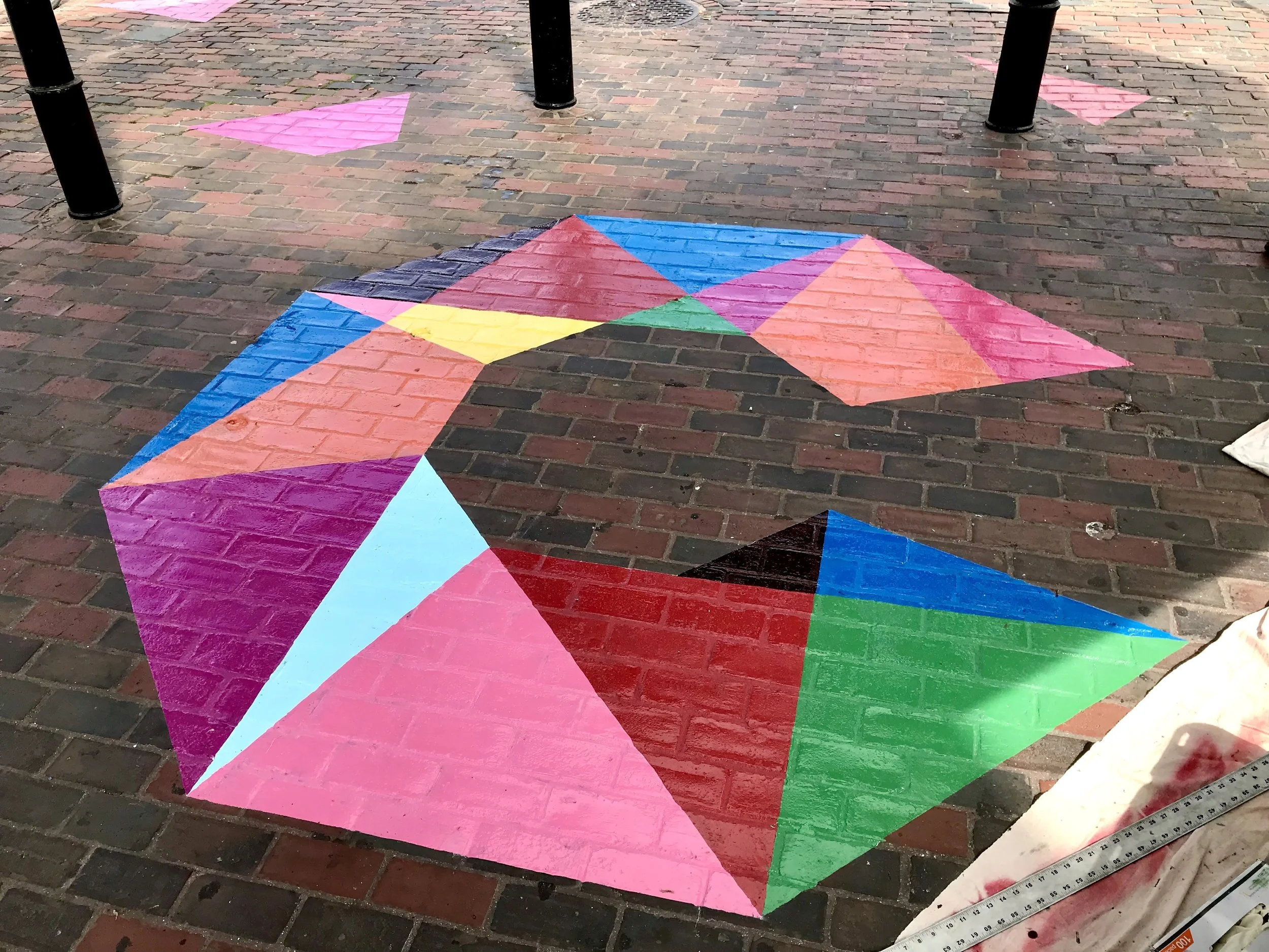 Colorful geometric street art painted on a brick sidewalk beside black bollards