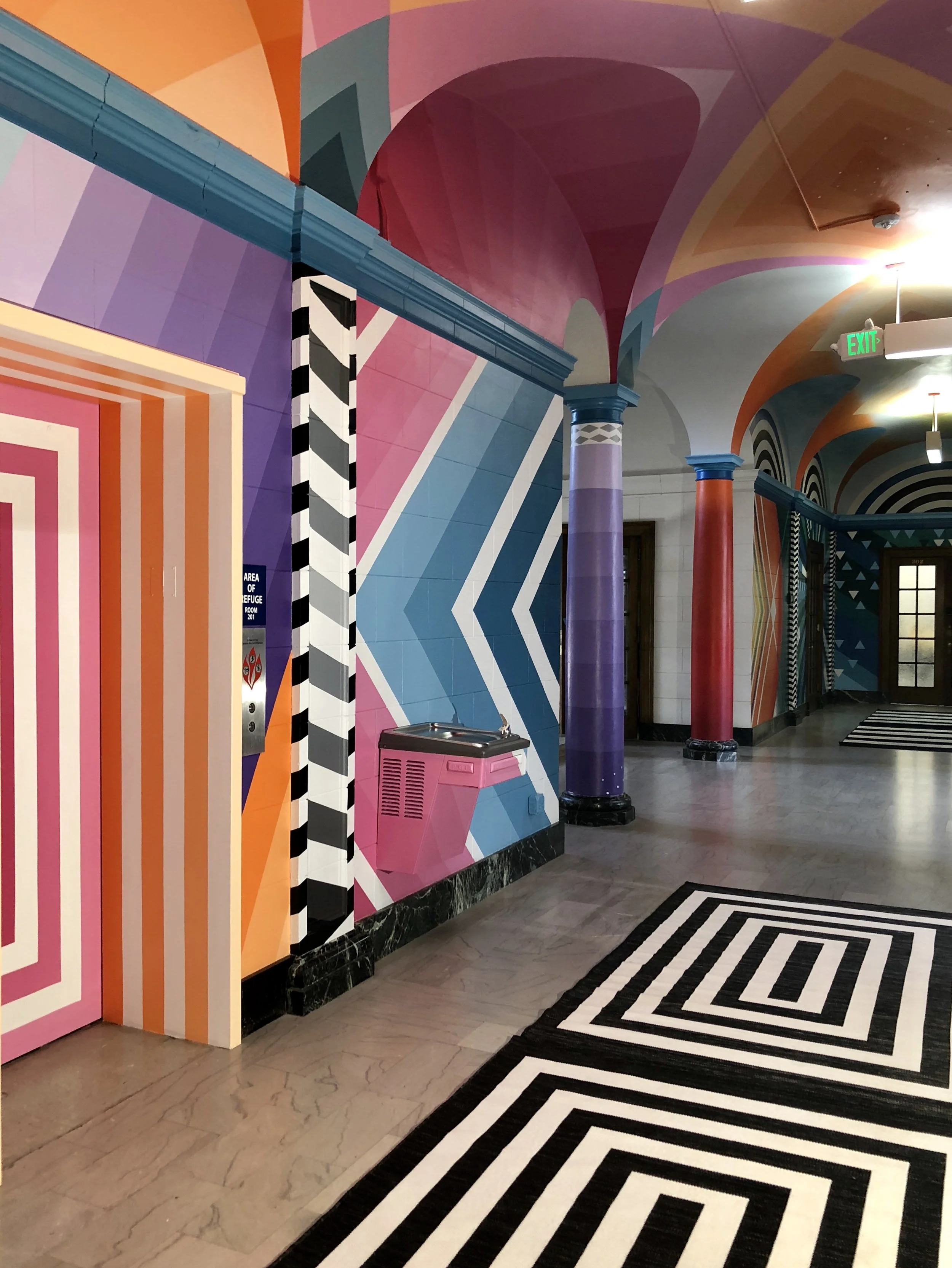 Colorful geometric hotel hallway with bold pink, orange, purple, and blue walls, columns, arched ceilings, patterned carpet, elevator, water fountain, and exit sign.