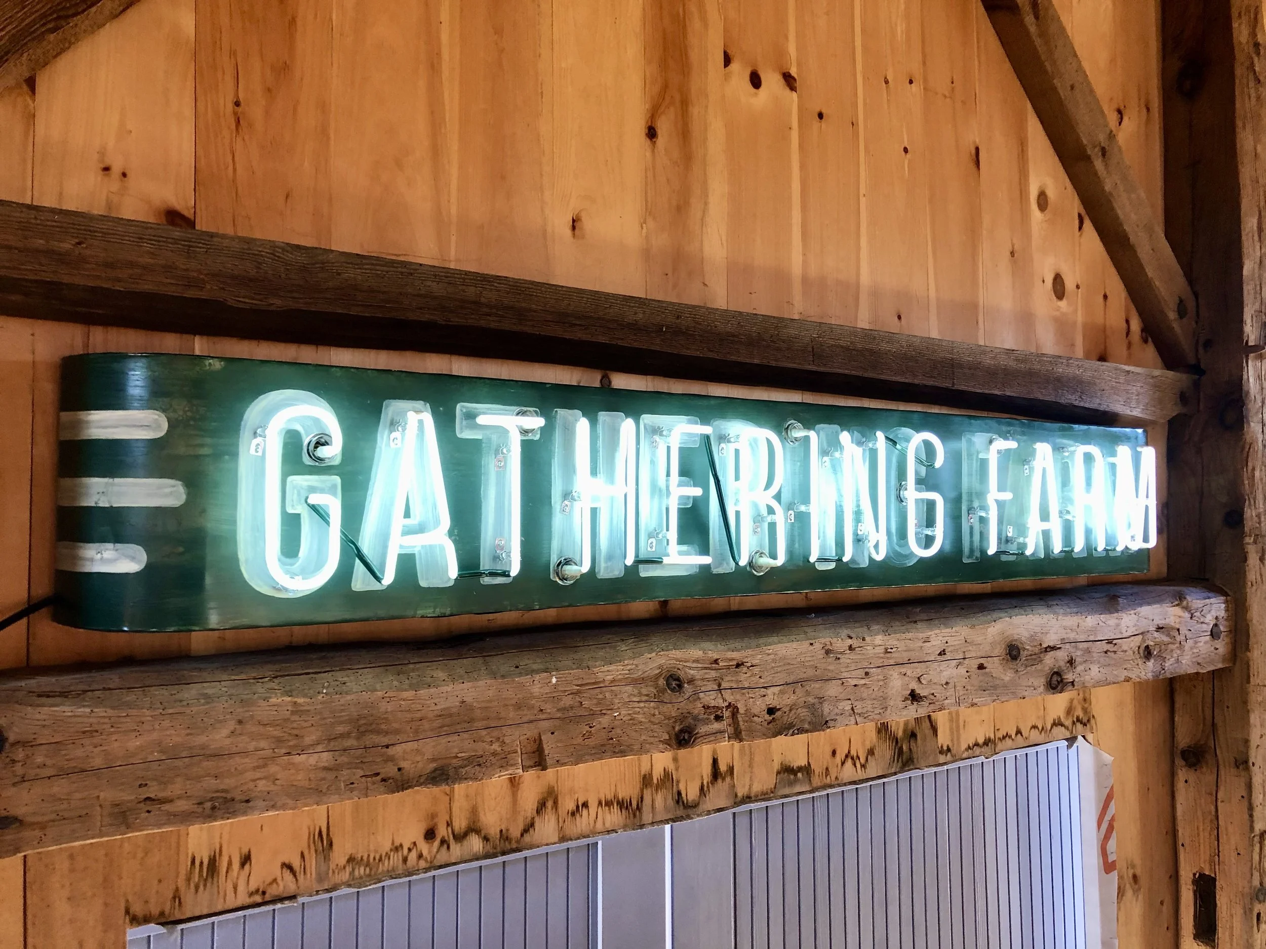 Glowing neon “GATHER HERE” sign mounted on a rustic wooden wall.