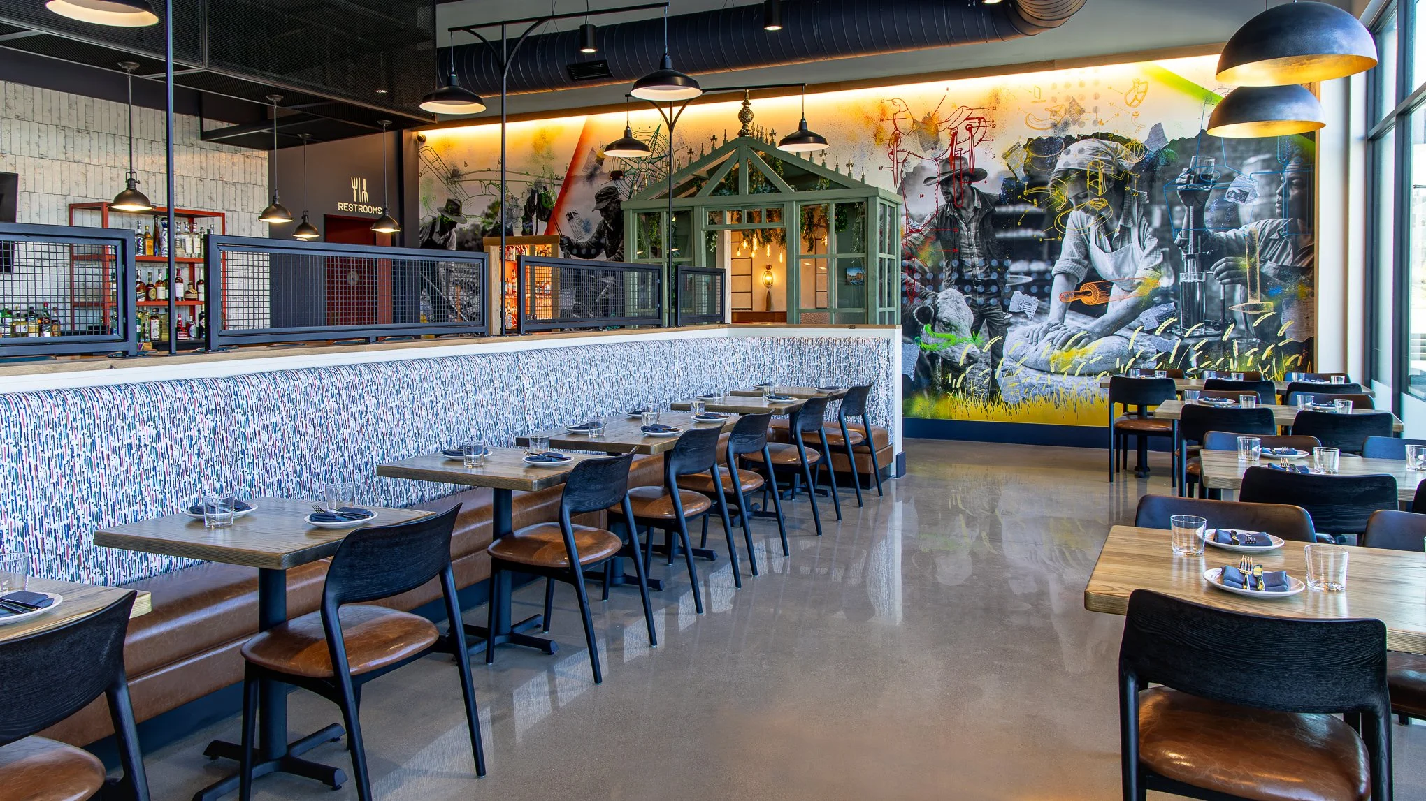 Modern restaurant interior with set dining tables, bar seating, pendant lights, and a large colorful mural