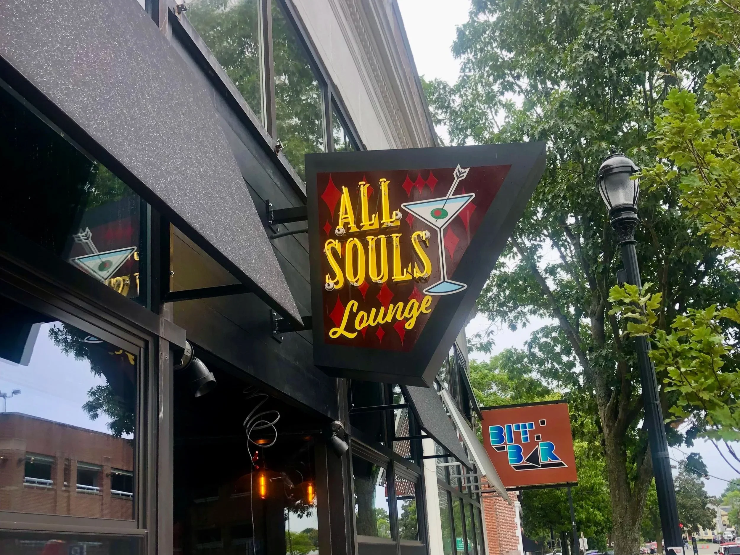 Exterior sign for All Souls Lounge with a martini glass logo outside a tree-lined storefront.