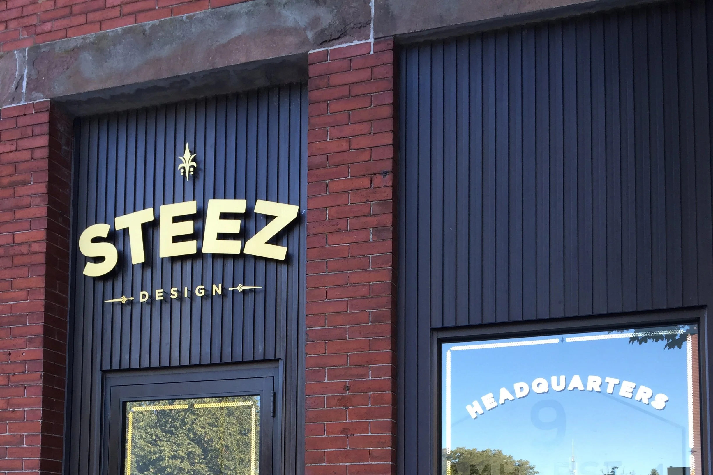 Brick building facade with a dark blue STEEZ DESIGN sign and a window labeled HEADQUARTERS.