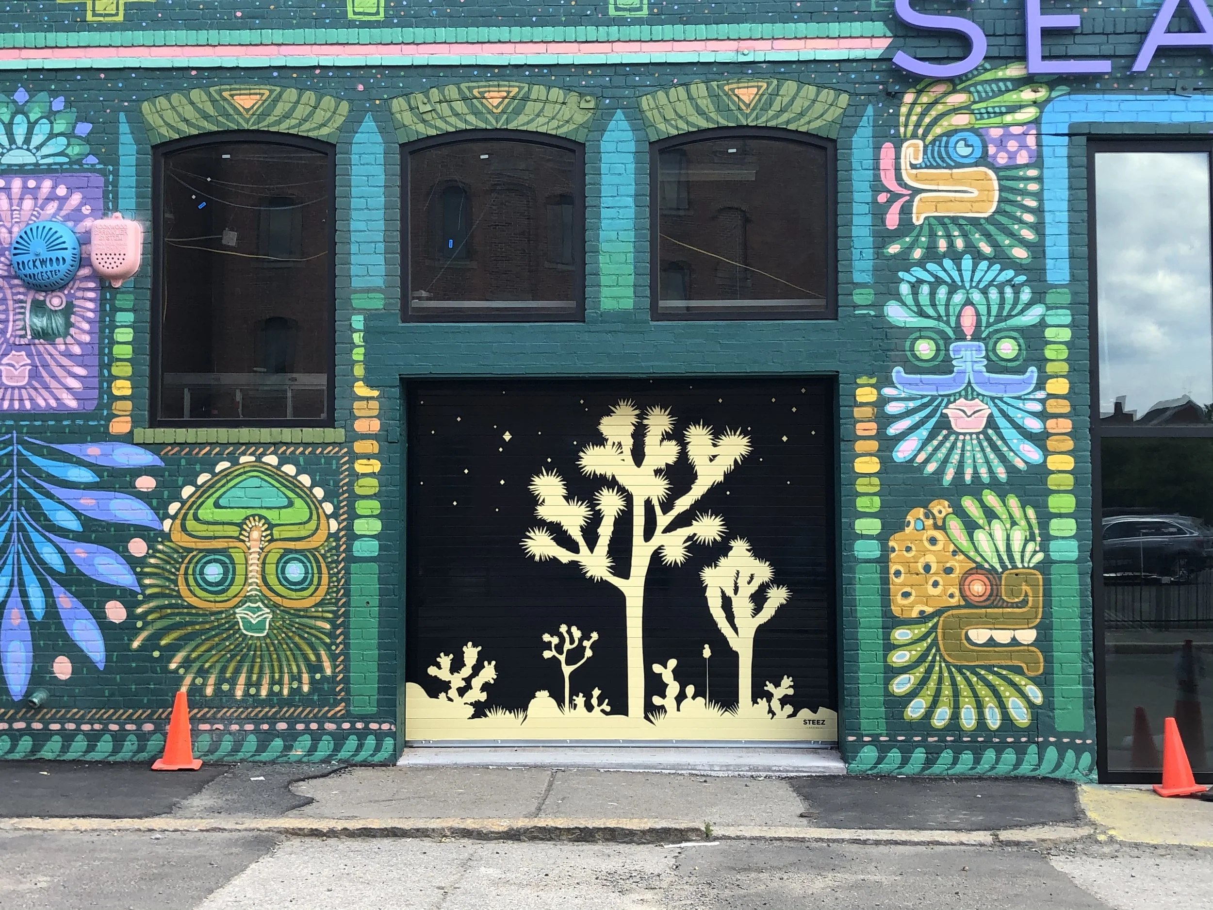 Colorful psychedelic mural on a brick storefront featuring stylized plants, flowers, insects, and a black garage door with desert silhouettes