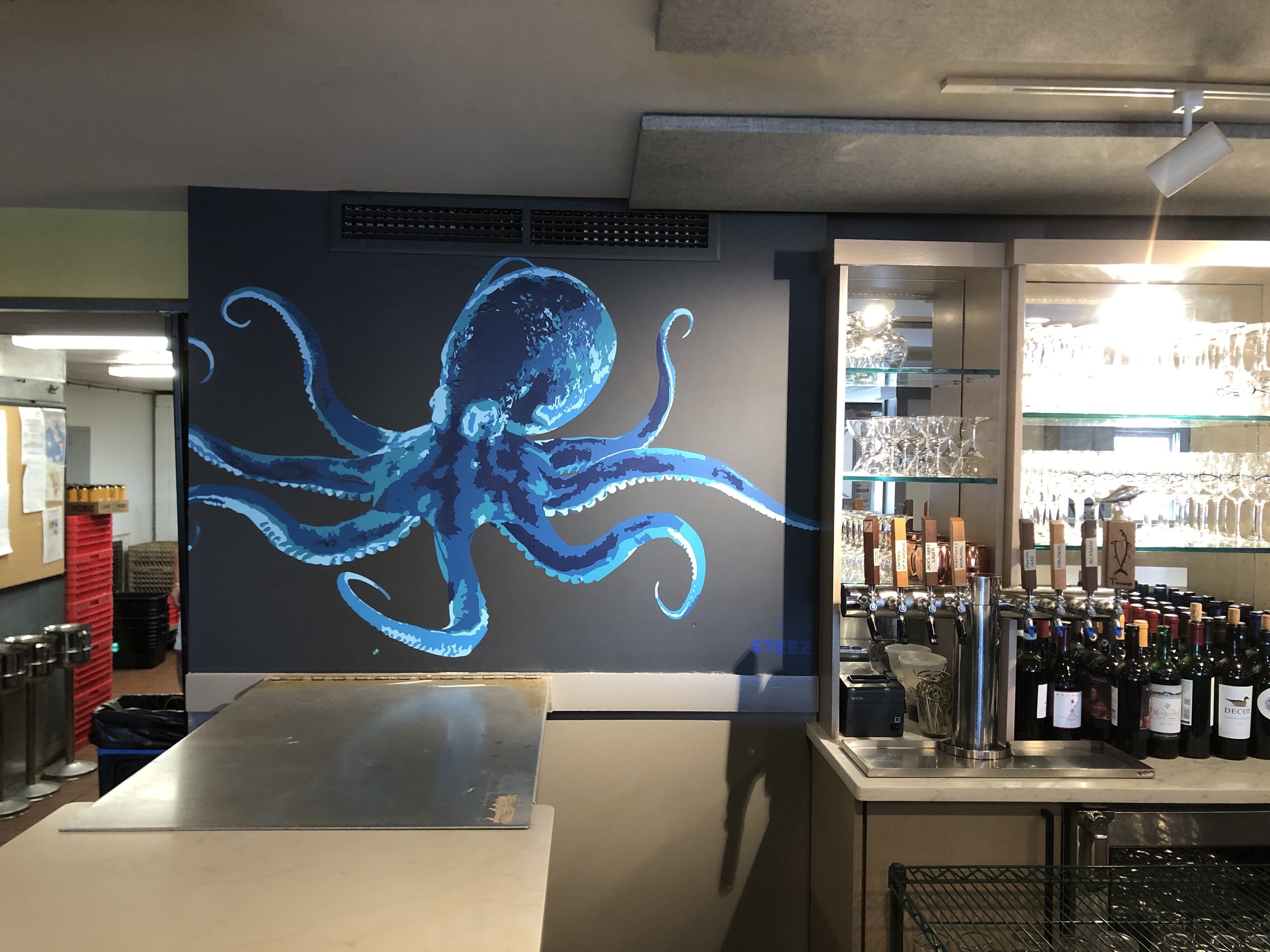 Restaurant bar with a large blue octopus mural, beer taps, wine bottles, and glassware