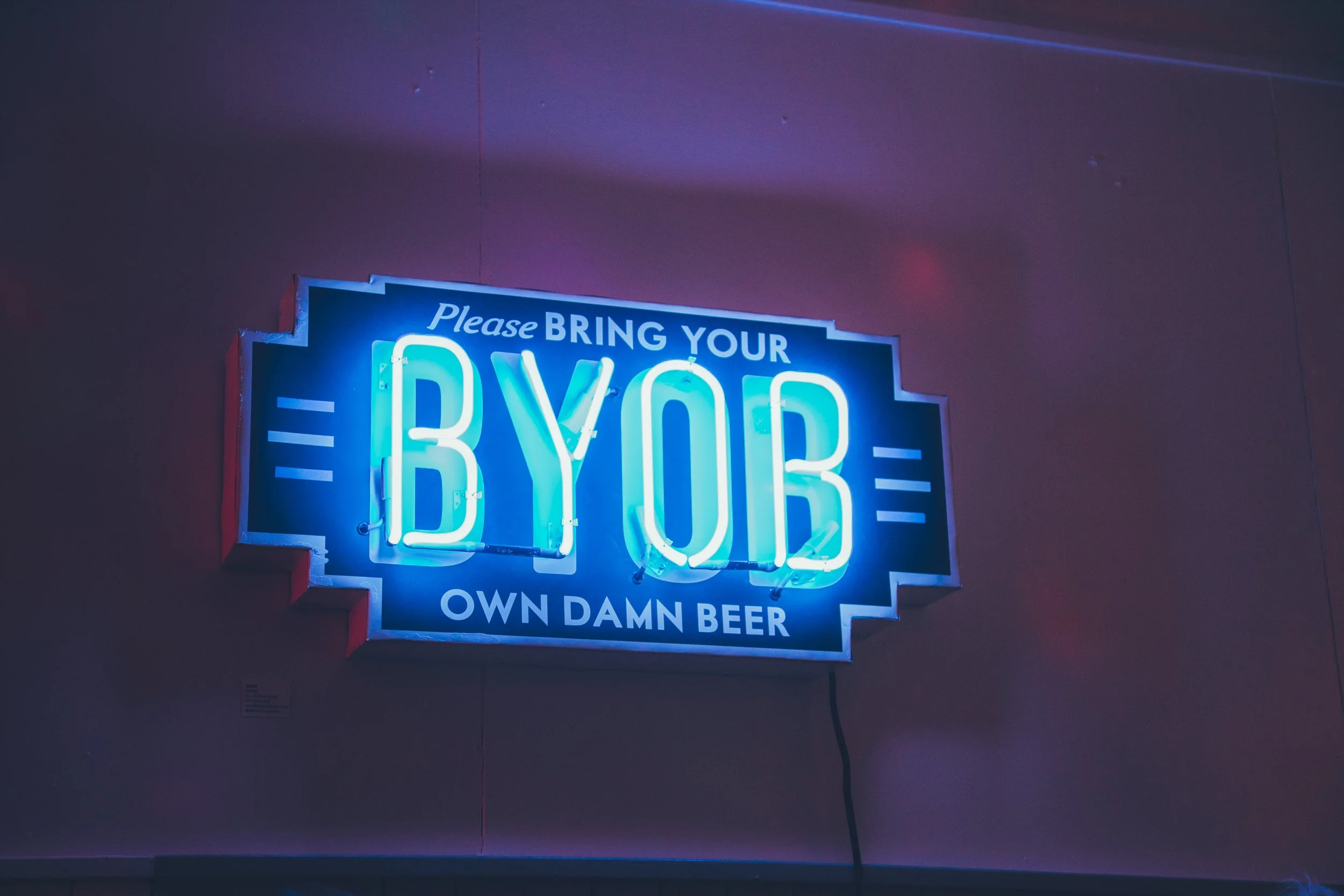 Blue neon sign reading “Please bring your own beer” on a dimly lit wall