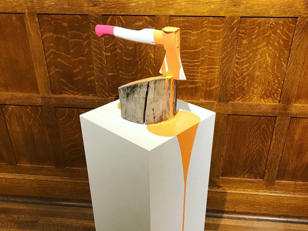 Decorative axe embedded in a log atop a white pedestal, with orange paint dripping down the front.