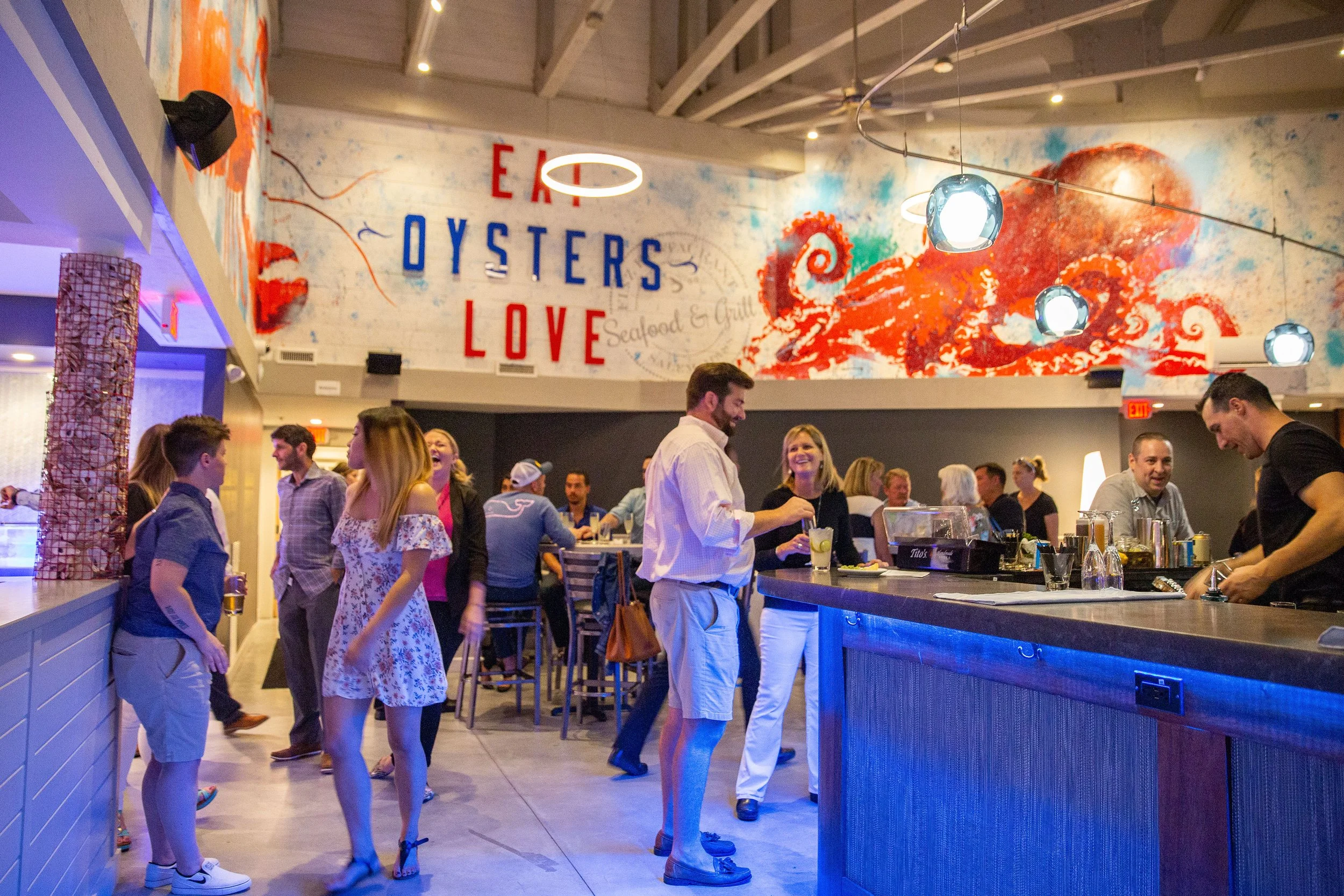 Busy seafood restaurant and bar with patrons, bartenders, colorful octopus mural, and "EAT OYSTERS LOVE" wall sign.