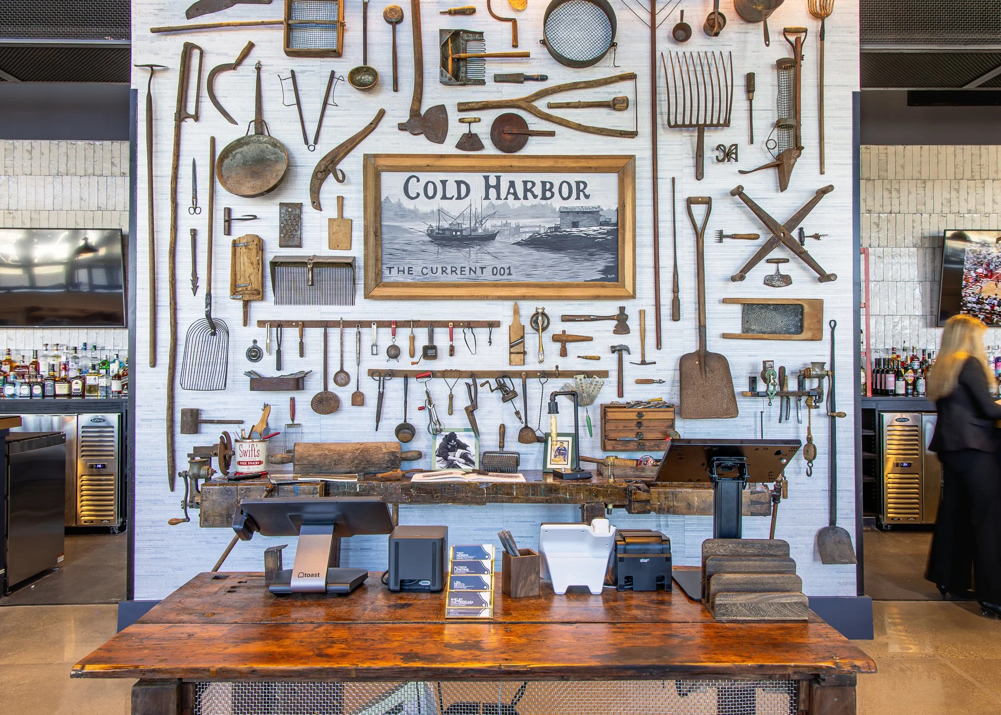 Restaurant display wall covered with vintage tools and kitchen implements, centered around a framed Cold Harbor harbor scene