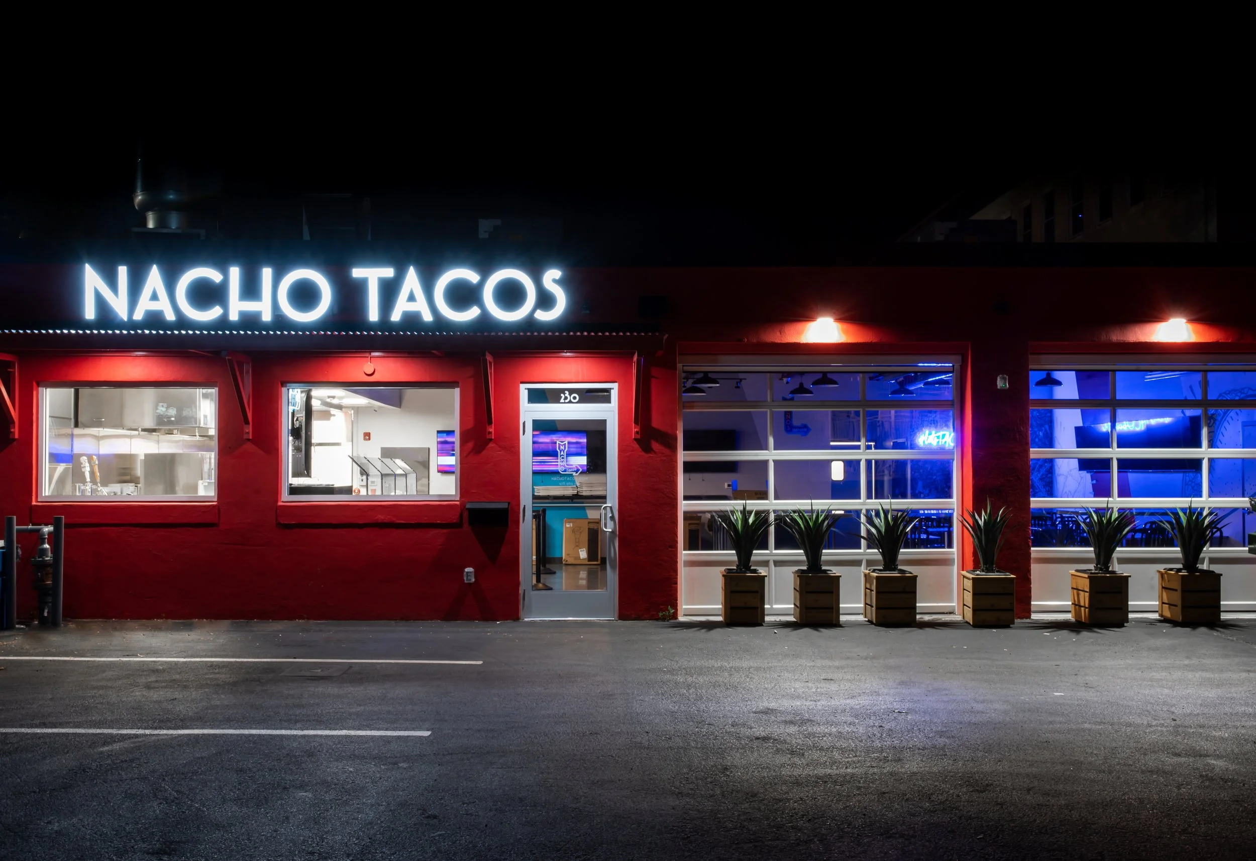 Nighttime exterior of Nacho Tacos restaurant with a glowing sign, red facade, blue-lit windows, and potted plants.