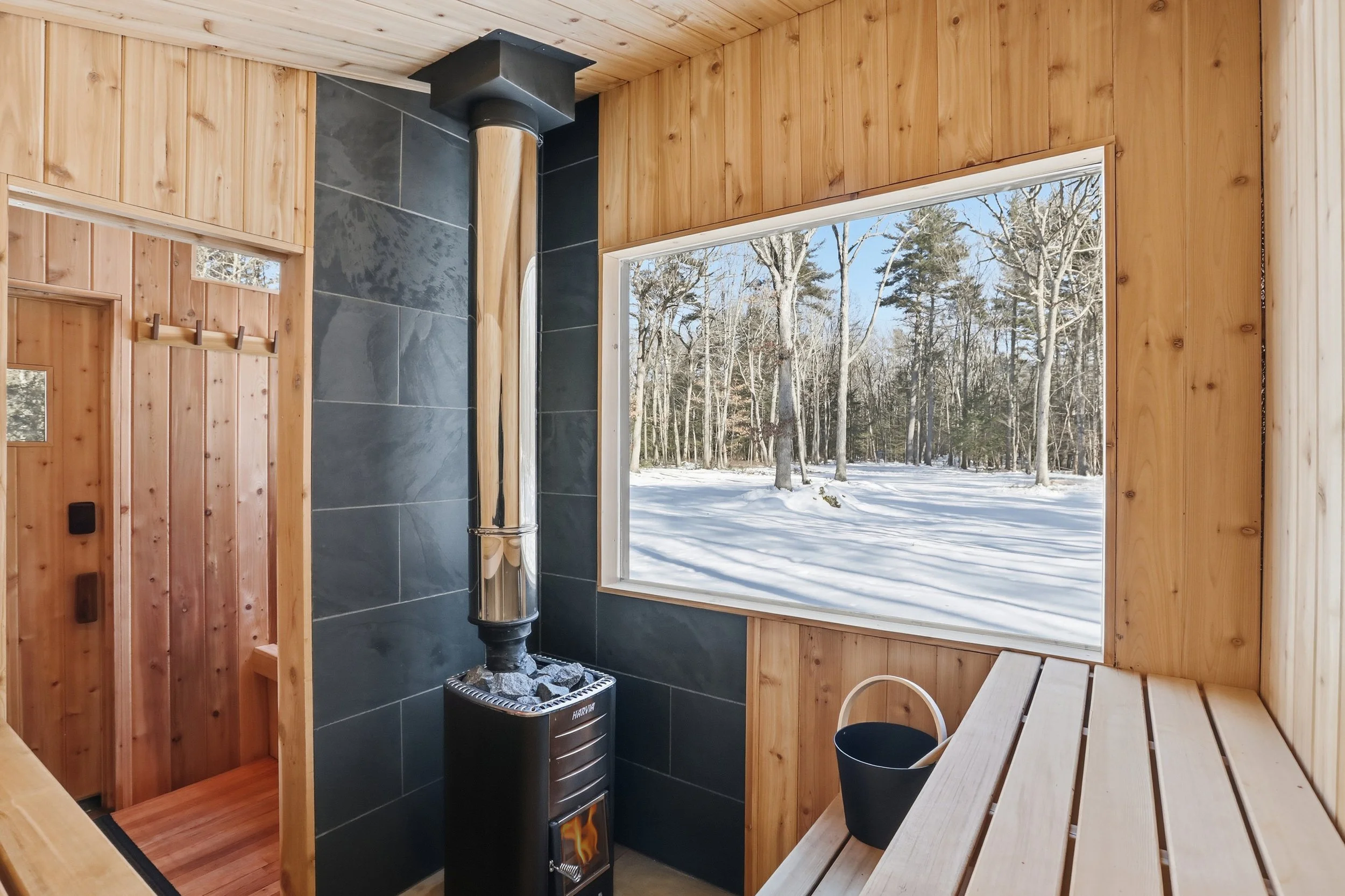 Wood-paneled sauna with a Harvia wood-burning stove, benches, and a large window overlooking a snowy forest.