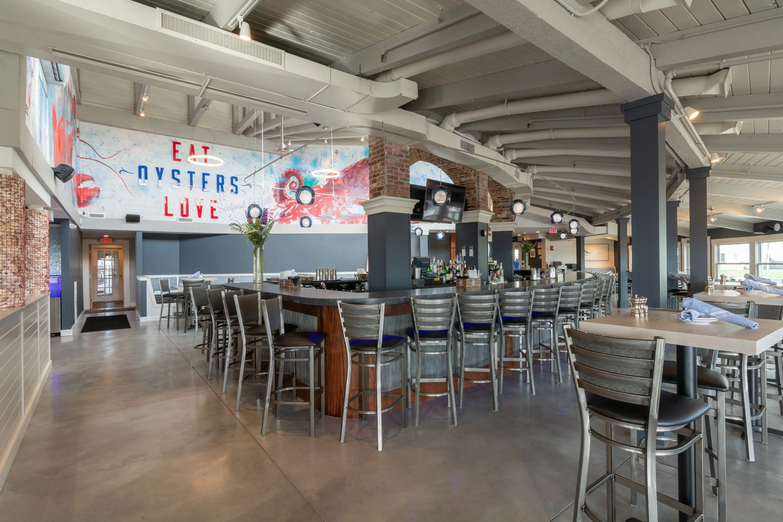Interior of a modern seafood restaurant with a large bar, high-top chairs, dining tables, exposed brick arches, and an “Eat Oysters Love” mural.