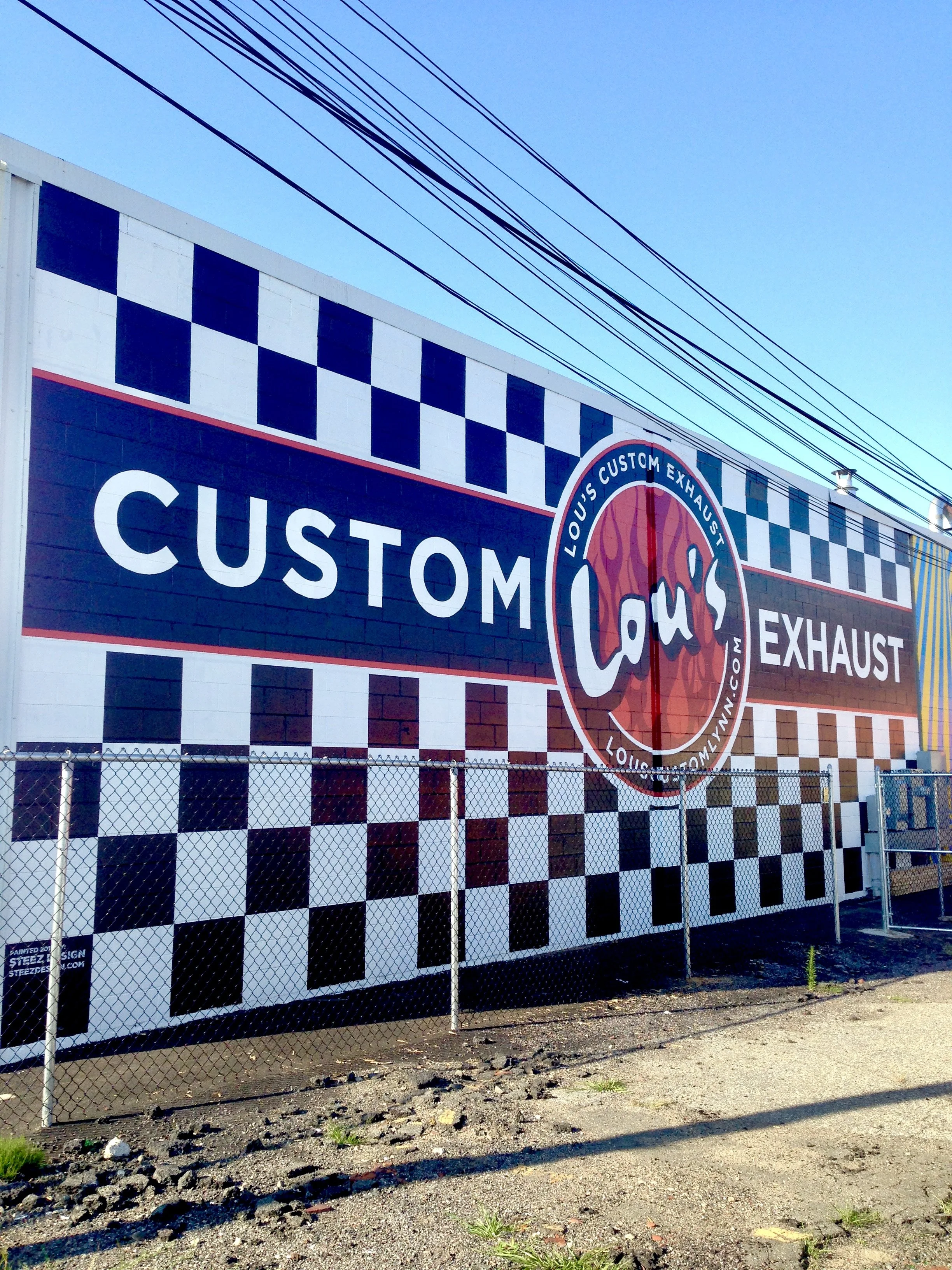 Exterior mural of Custom Exhaust automotive shop with checkerboard pattern, colorful exhaust graphic, chain-link fence, and overhead utility wires.