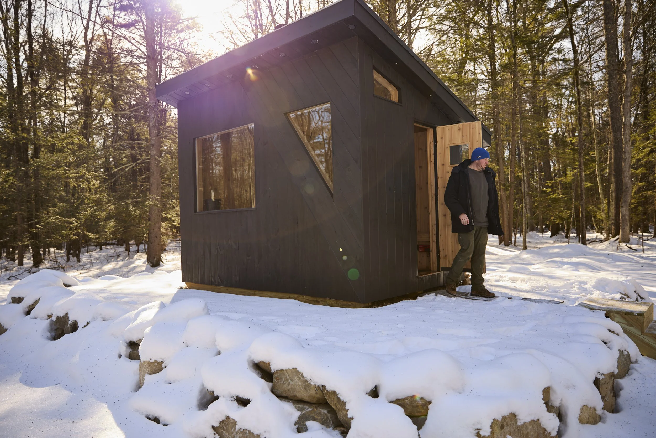Modern black tiny cabin with a wooden door in a snowy forest, with a man standing outside