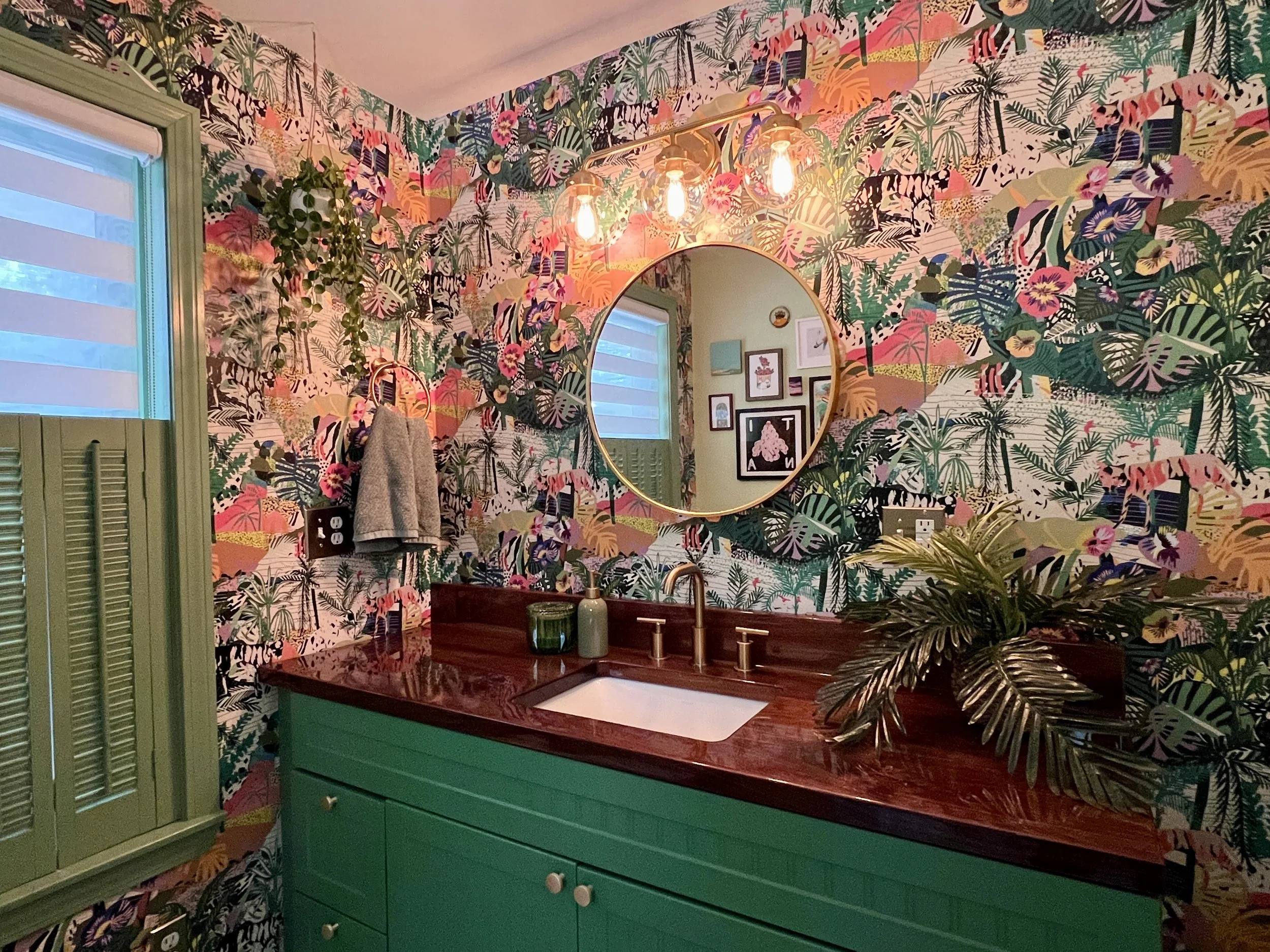 Colorful tropical botanical wallpapered bathroom with green vanity, burgundy countertop, vessel sink, round gold-framed mirror, pendant lights, and lush plants