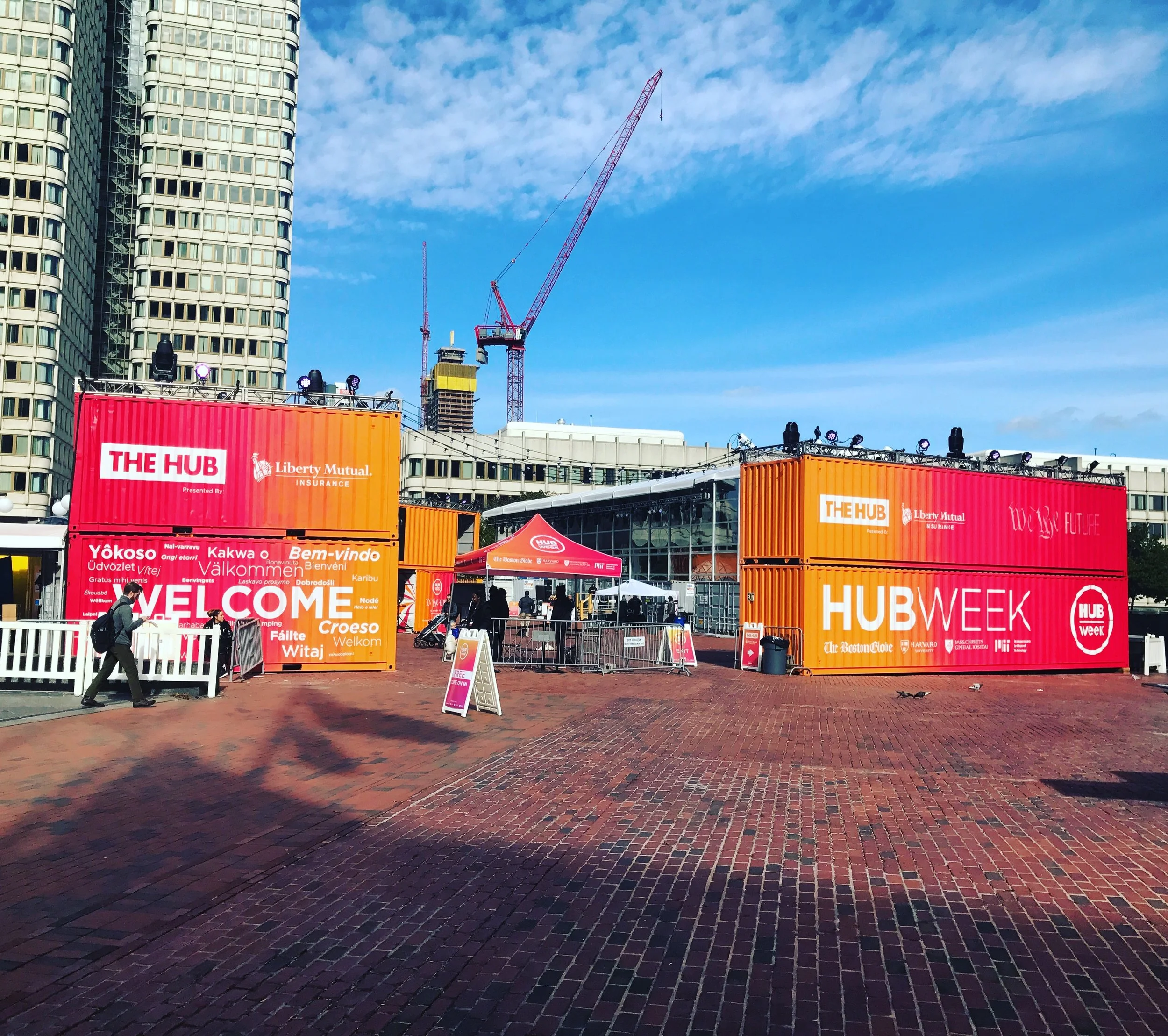 HubWeek event venue with colorful shipping-container booths, stage lighting, construction cranes, and a city office building under a blue sky