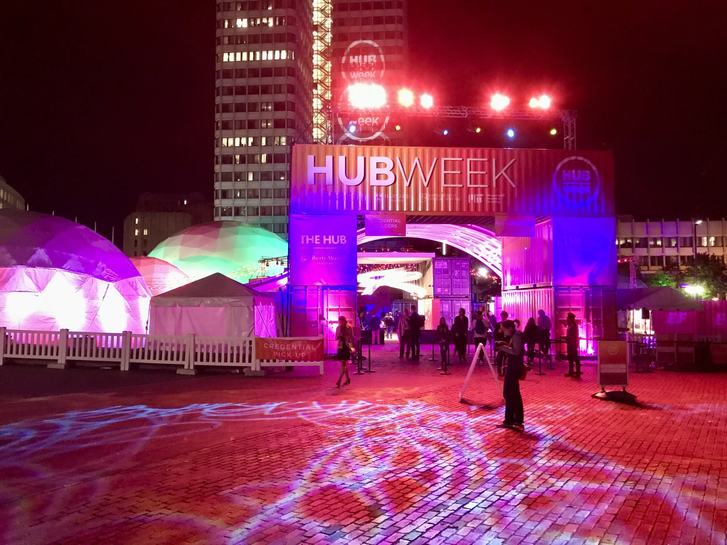 Nighttime HubWeek event entrance illuminated with colorful lights, shipping containers, tents, and attendees in an urban plaza