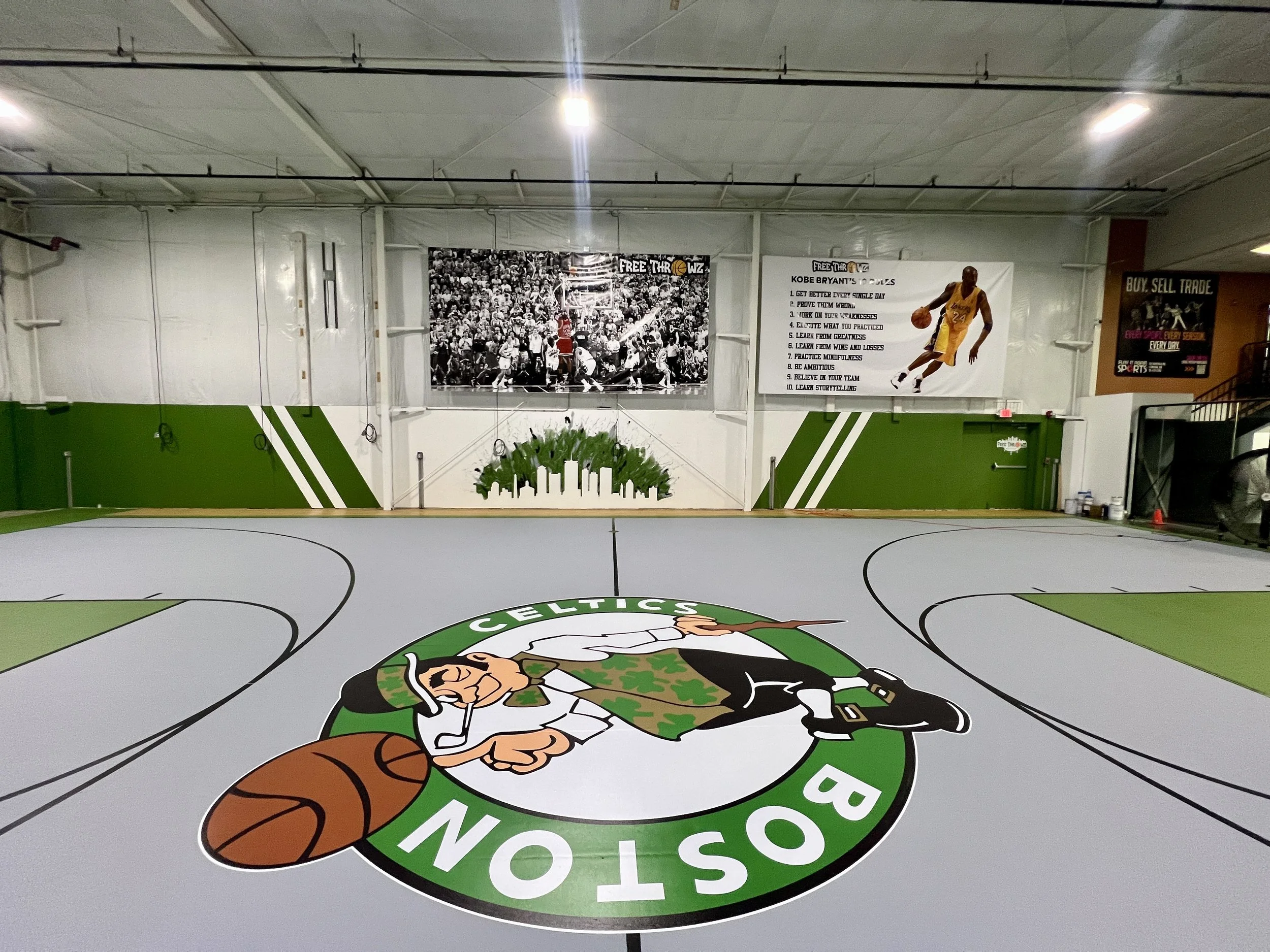 Indoor basketball court with a Boston Celtics-style center logo, green-and-white walls, and Kobe Bryant basketball posters.