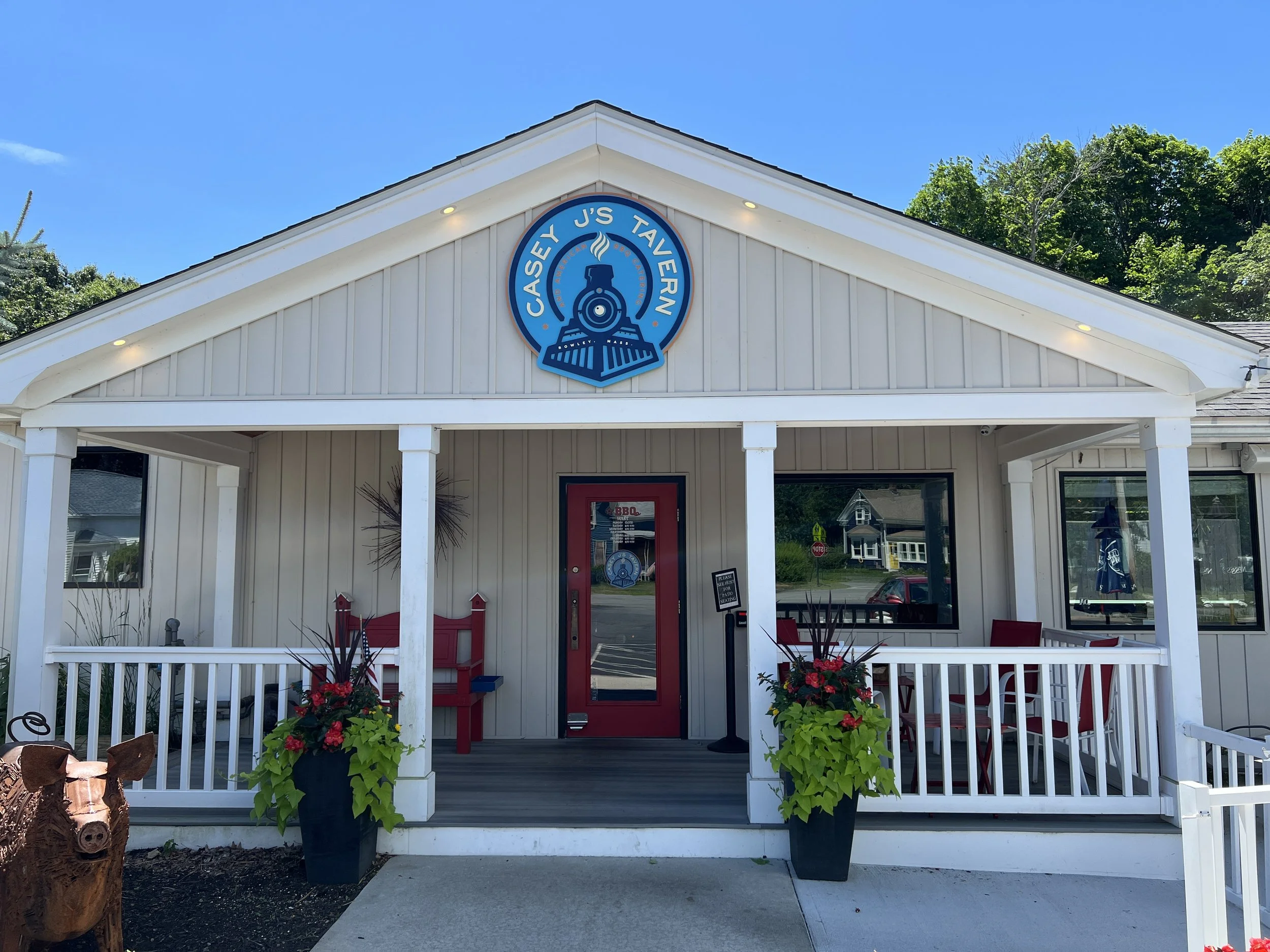 Exterior of Casey J's Tavern in Rowley, Massachusetts, with a white porch, red door, restaurant sign, and flower planters.