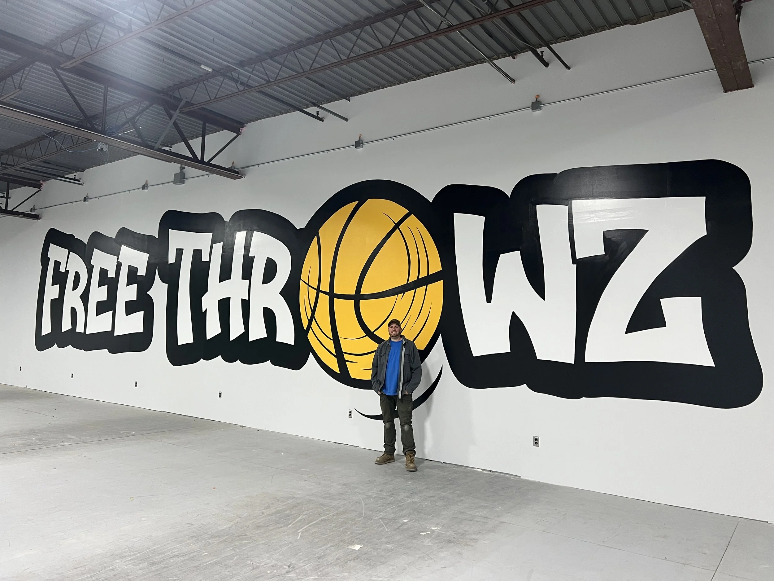 Man standing in an industrial gym beneath a large “FREE THROW” basketball mural with a yellow basketball graphic.