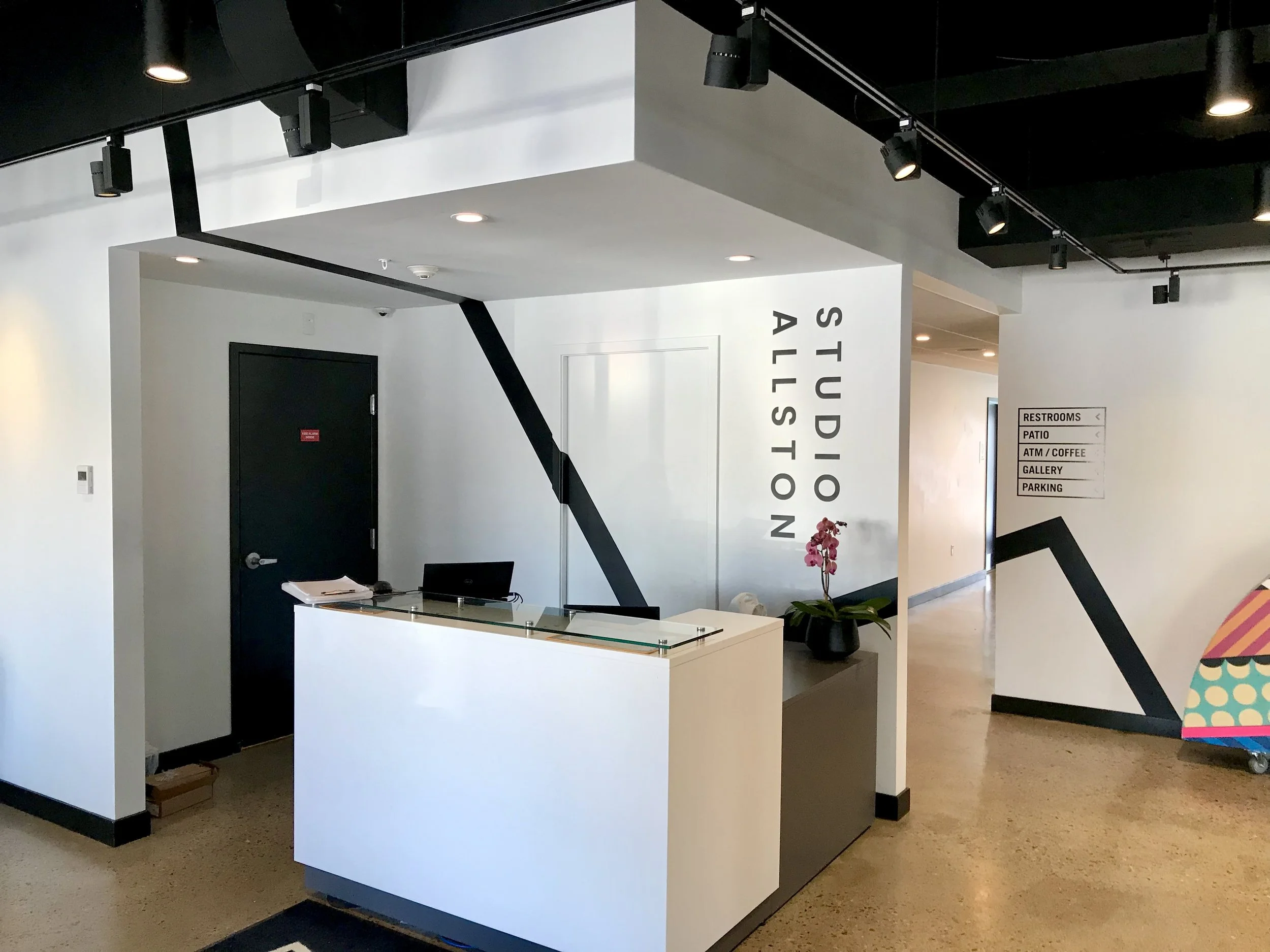 Modern gallery lobby with a white reception desk, orchid, black geometric accents, and signs for restrooms, patio, ATM, coffee, gallery, and parking.