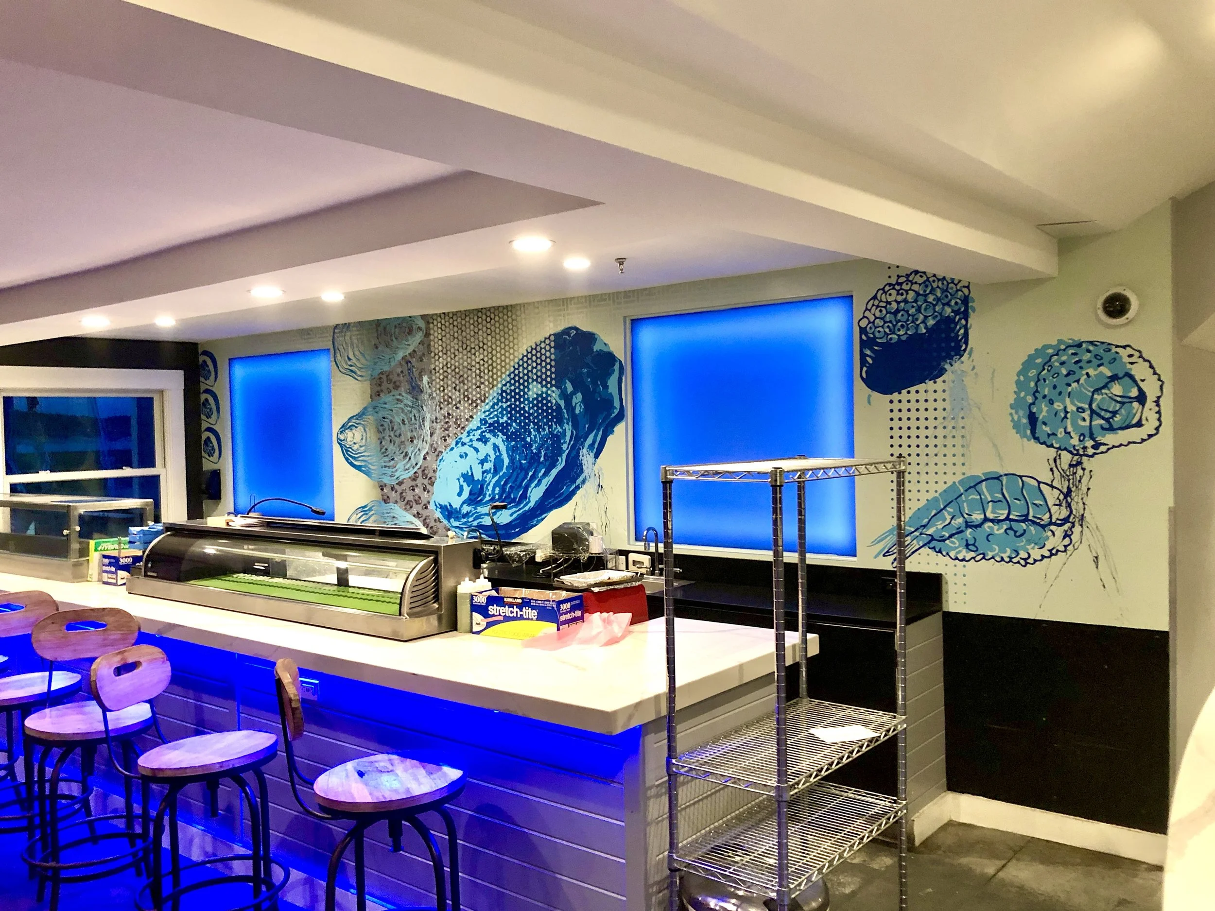 Modern sushi bar with blue lighting, sushi display case, stools, metal shelving, and ocean-themed wall murals