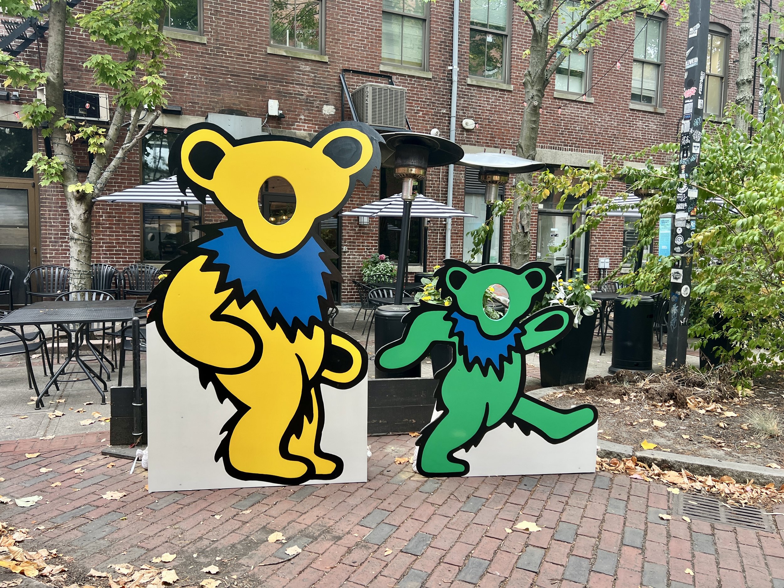 Large yellow and smaller green Grateful Dead dancing bear cutouts outside a brick building with an outdoor café patio.