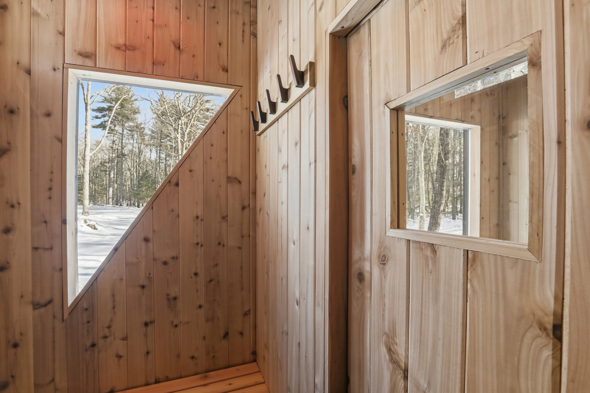 Wood-paneled cabin entryway with coat hooks, bench, windows, and a snowy forest outside