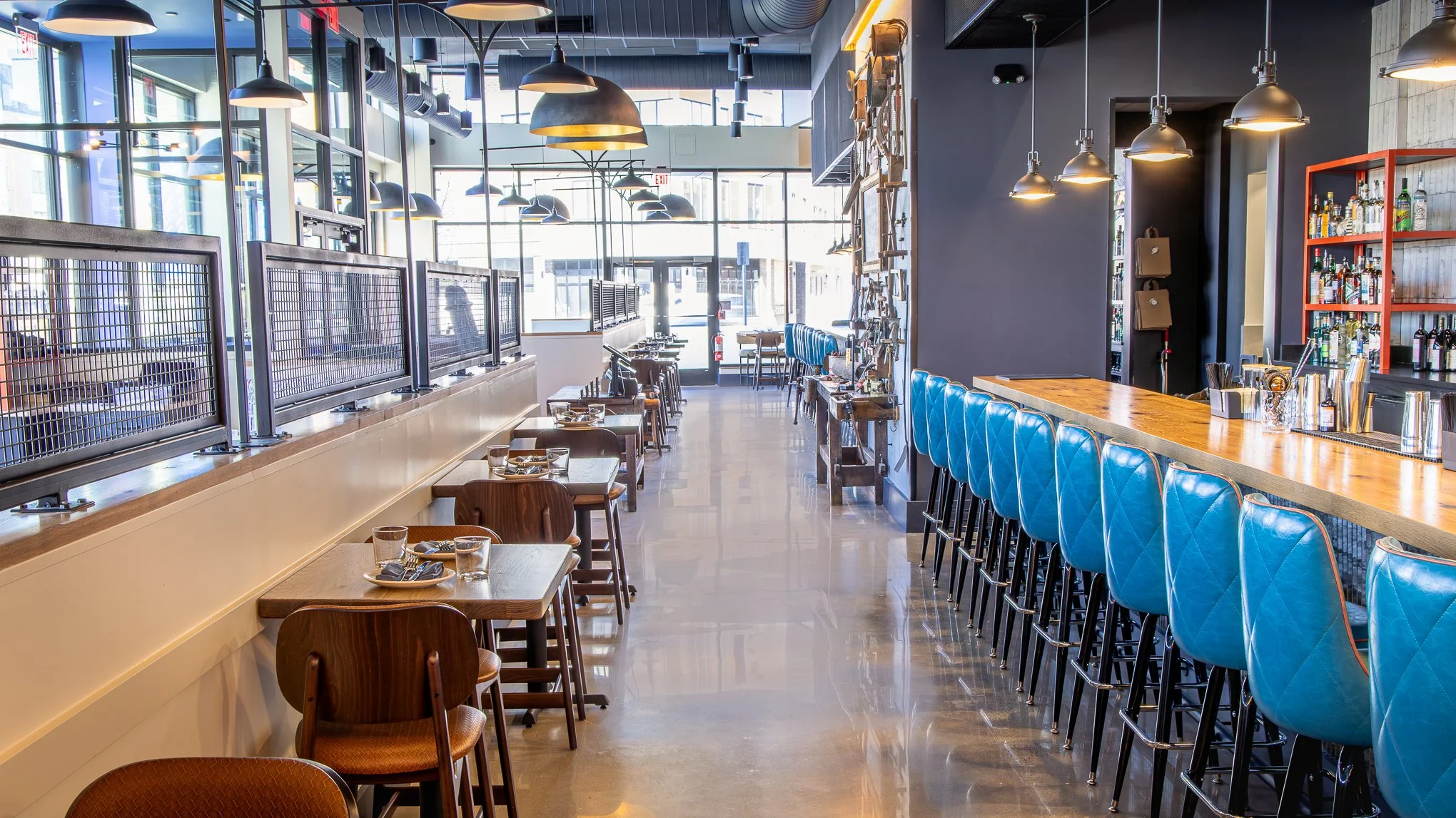Bright modern restaurant interior with blue bar stools, wood tables, pendant lighting, and a stocked liquor bar.