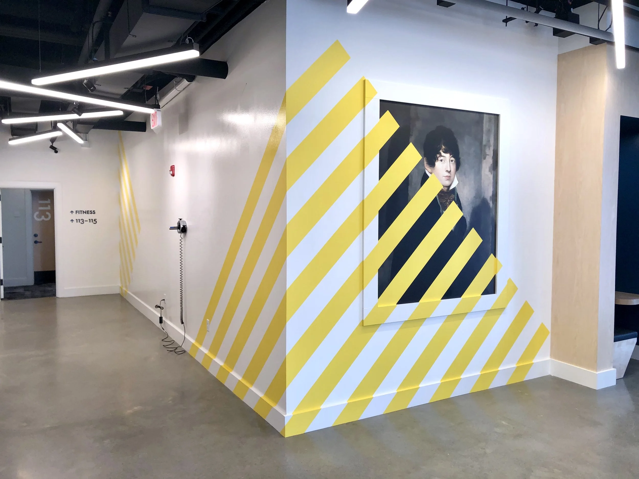 Modern hallway with yellow diagonal wall stripes, a portrait of a 19th-century man, and signs directing visitors to fitness rooms 113–115.
