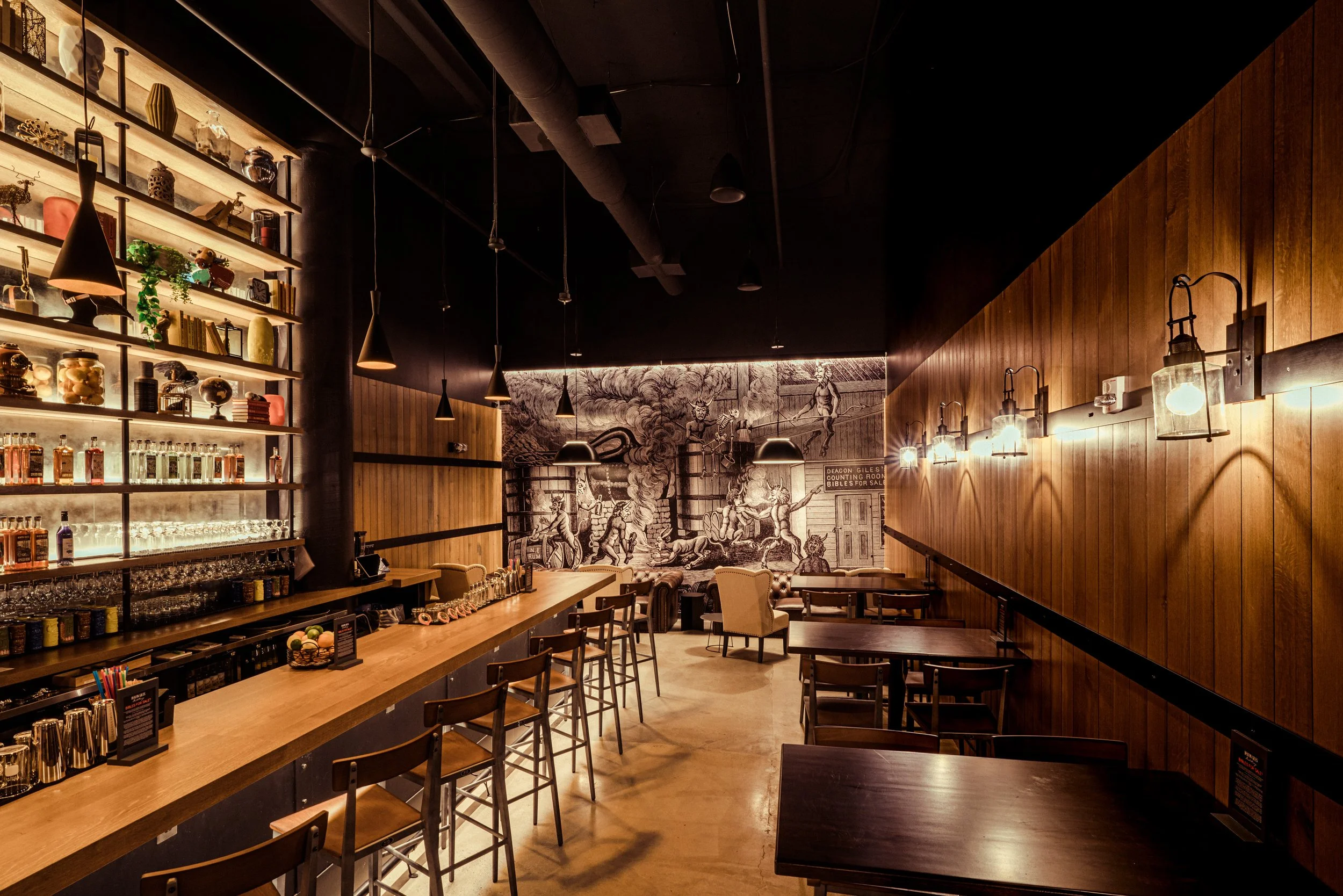 Warmly lit cocktail bar interior with liquor shelves, wooden paneling, pendant lights, lounge seating, and a large vintage mural