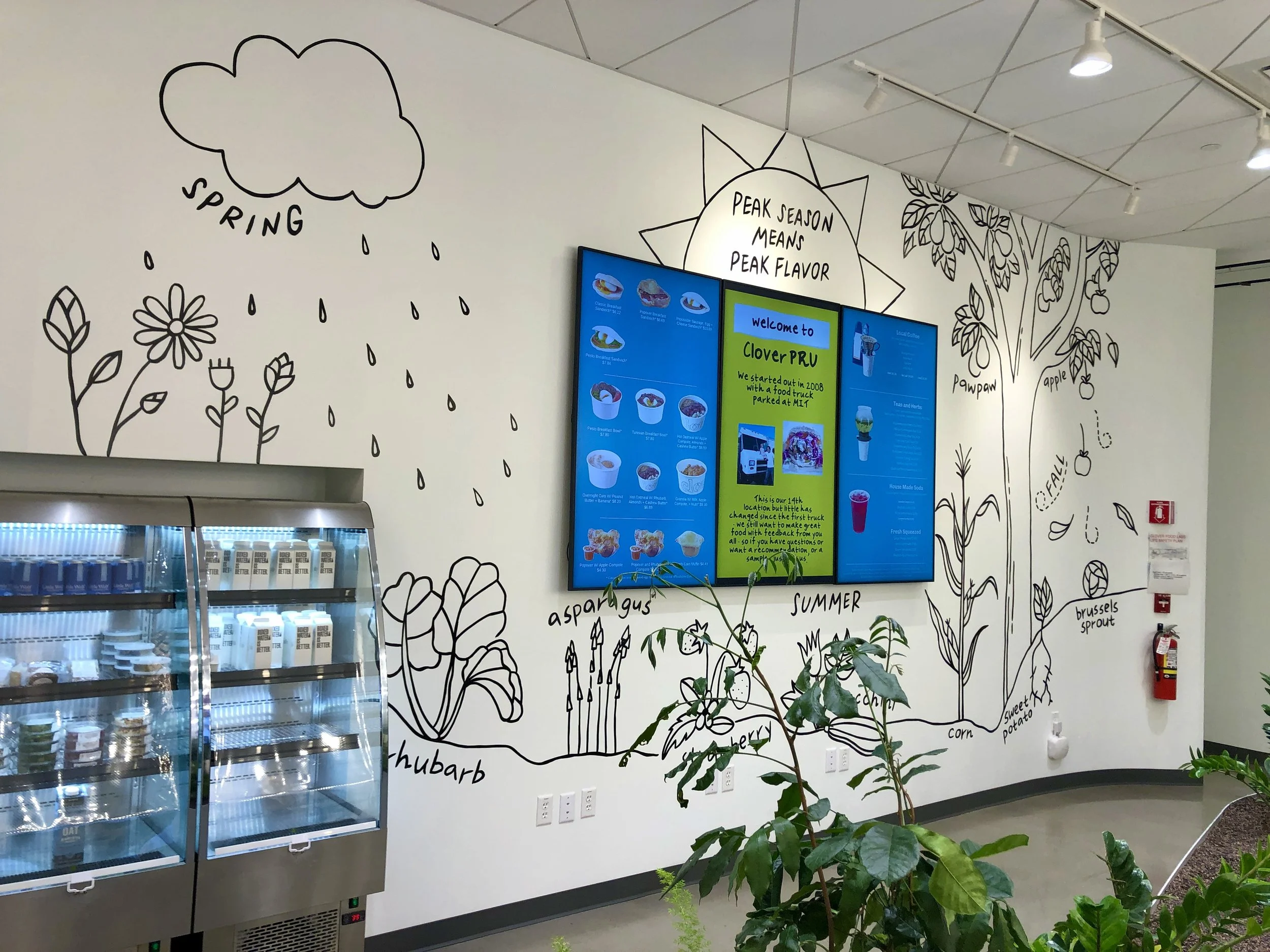 Clover Food Lab cafe interior with seasonal produce mural, digital menu screens, refrigerated food display, and indoor plants