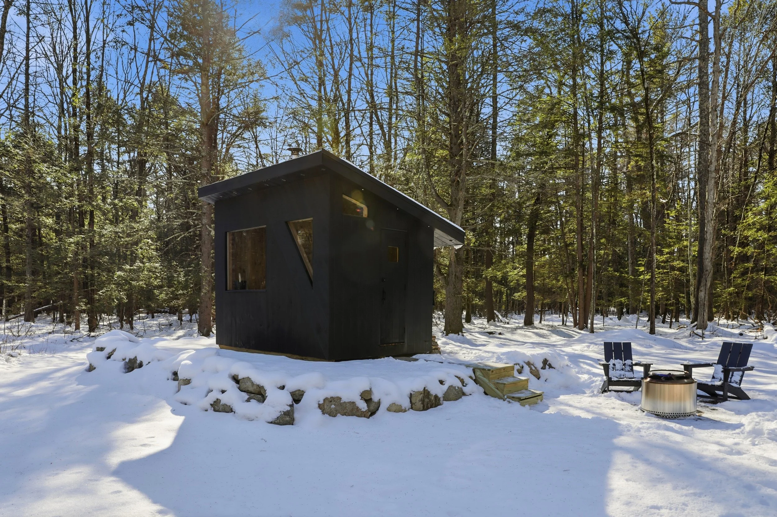 Modern black cabin surrounded by snowy forest, with Adirondack chairs and a fire pit outside