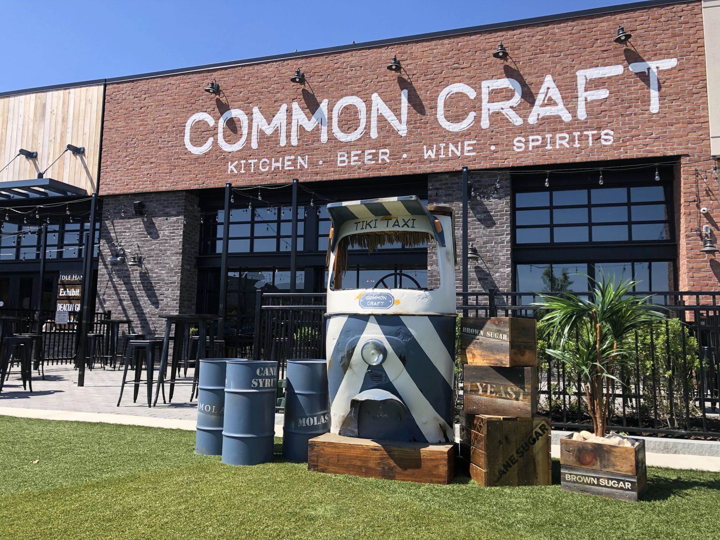 Common Craft Kitchen, Beer, Wine & Spirits restaurant exterior with a tiki taxi display, barrel tables, and outdoor patio
