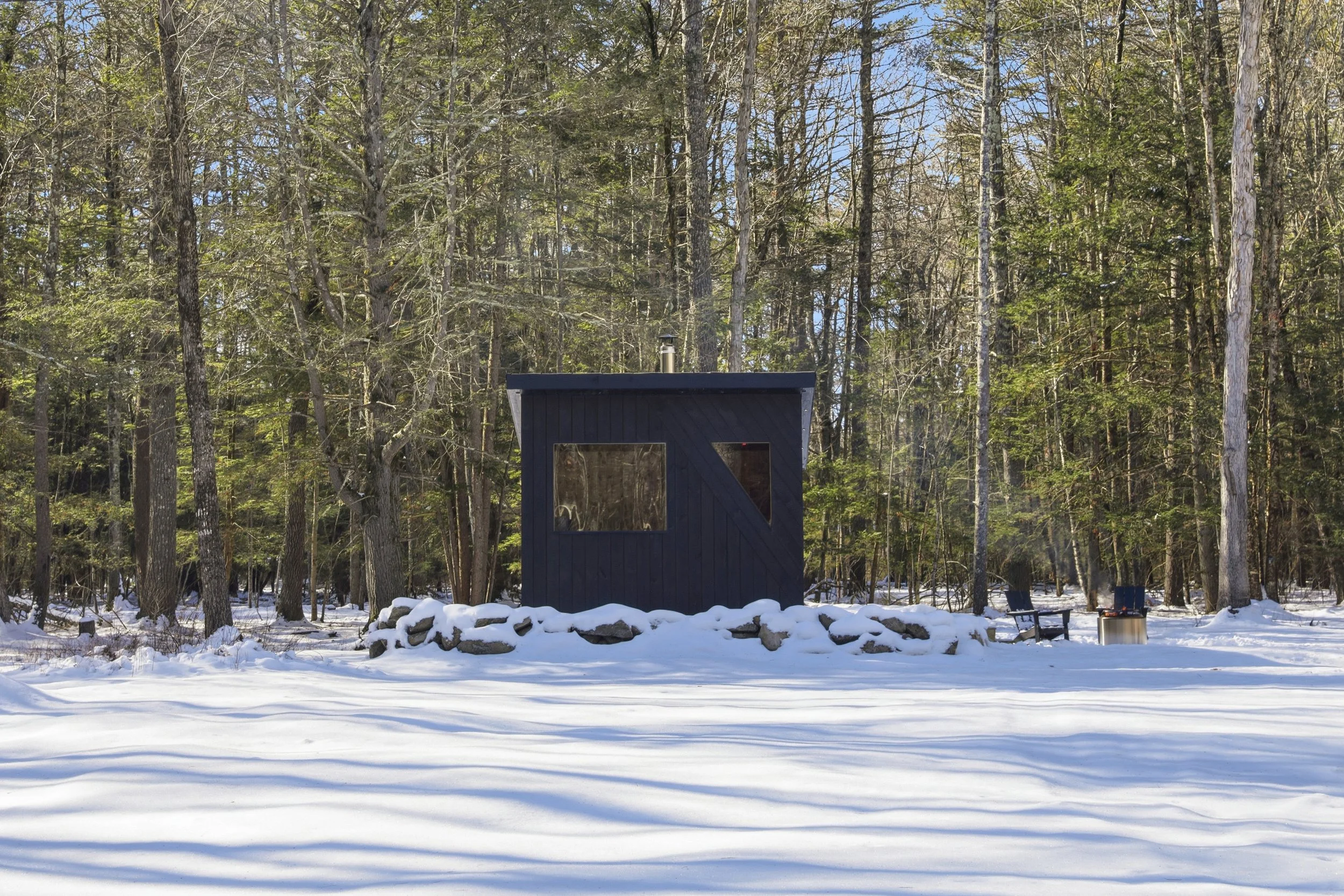 Small dark cabin with a chimney in a snowy forest, surrounded by trees and outdoor seating.