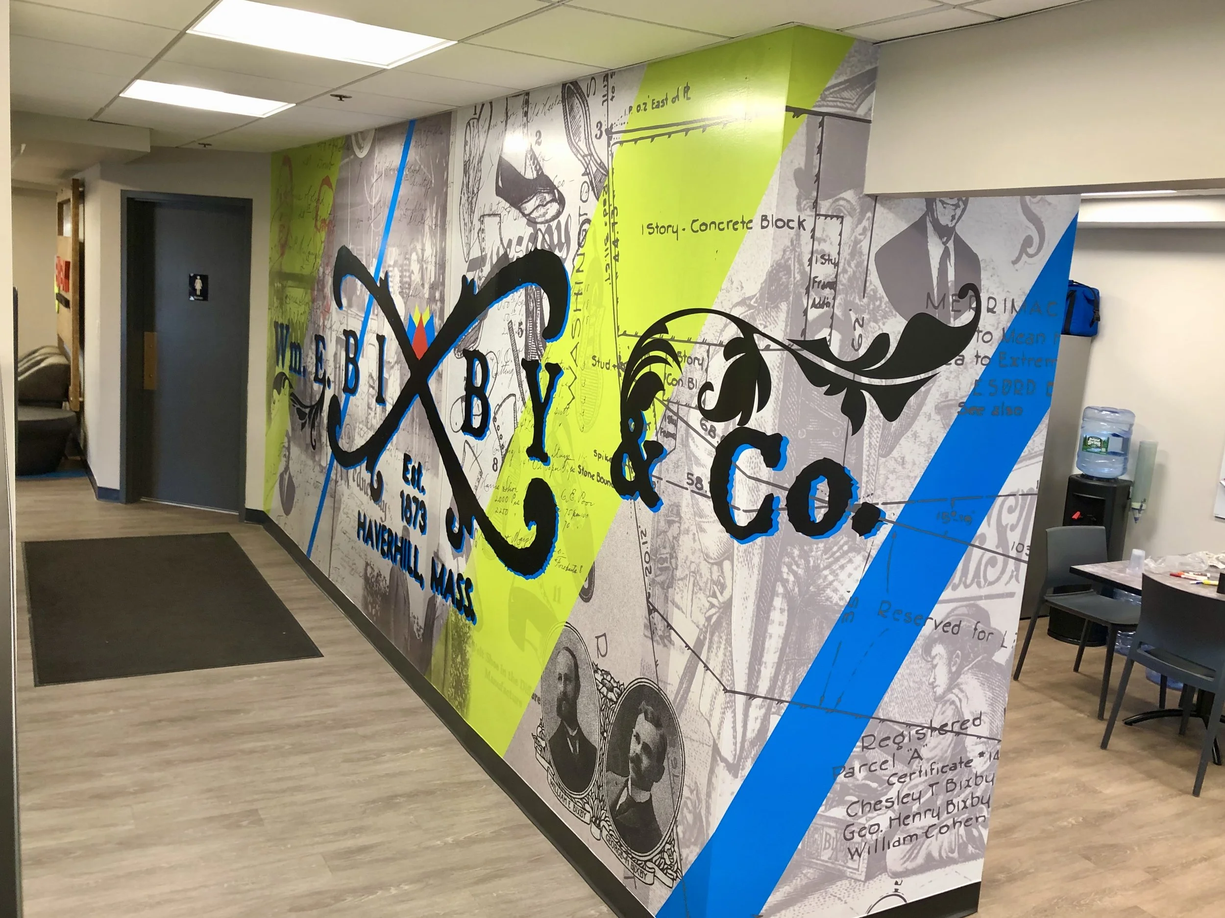 Colorful graffiti-style historical mural in an office hallway with a restroom, seating area, and water cooler.