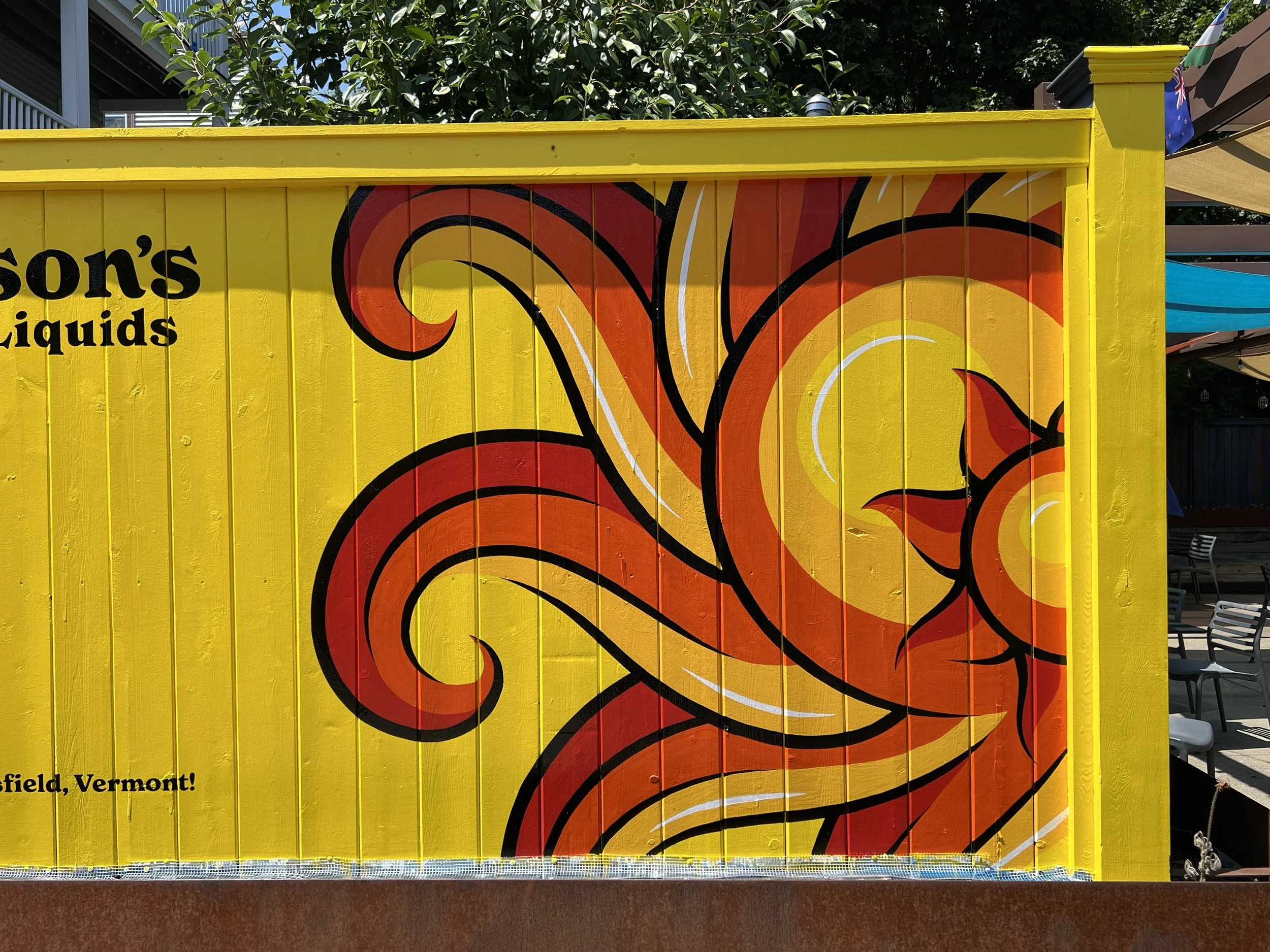 Bright yellow wooden mural with large abstract orange and red floral artwork outside a cafe, with partial text and outdoor seating.
