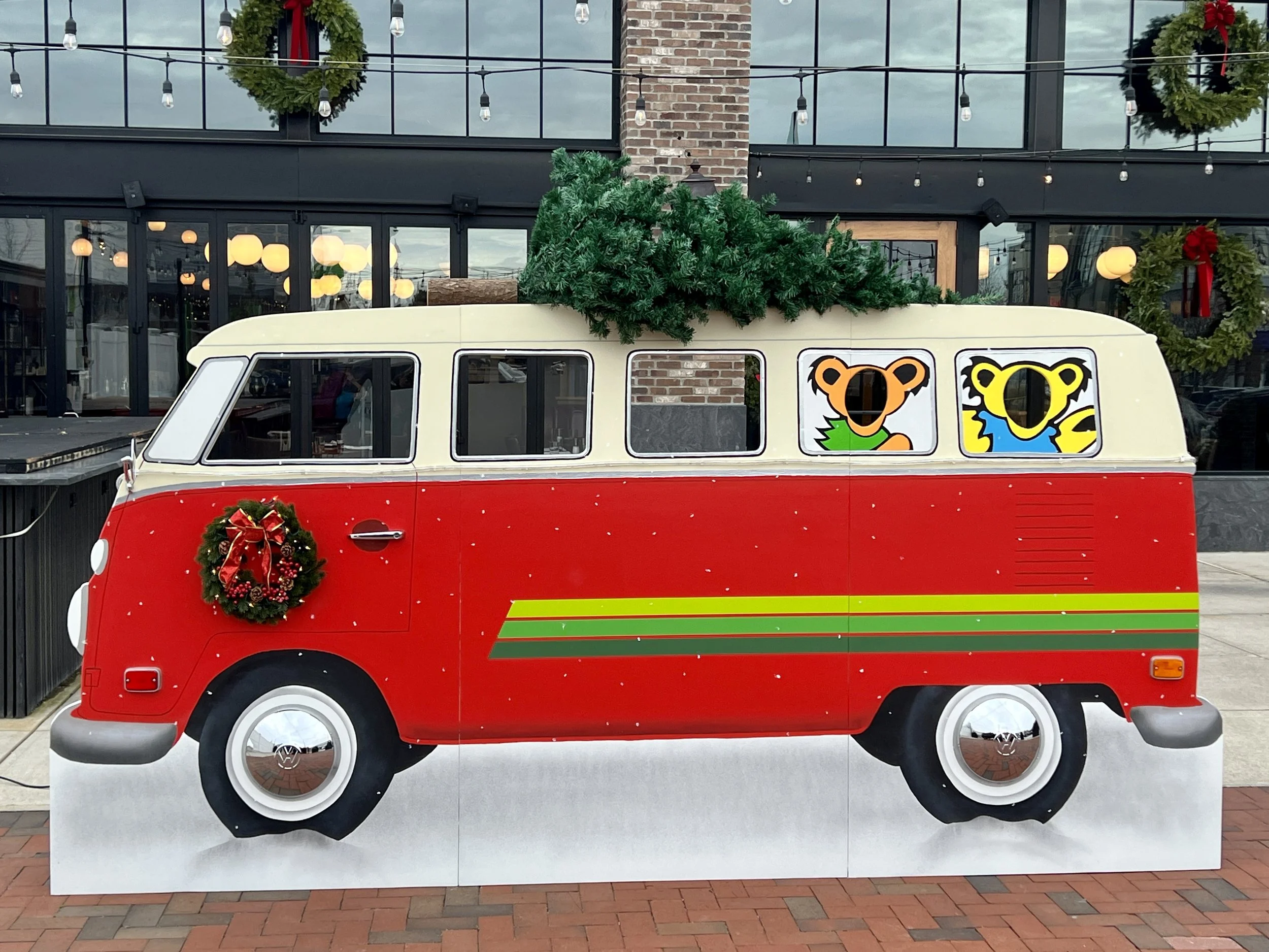 Holiday-decorated red-and-cream Volkswagen bus with wreath, Christmas tree, and colorful bear decals outside a restaurant