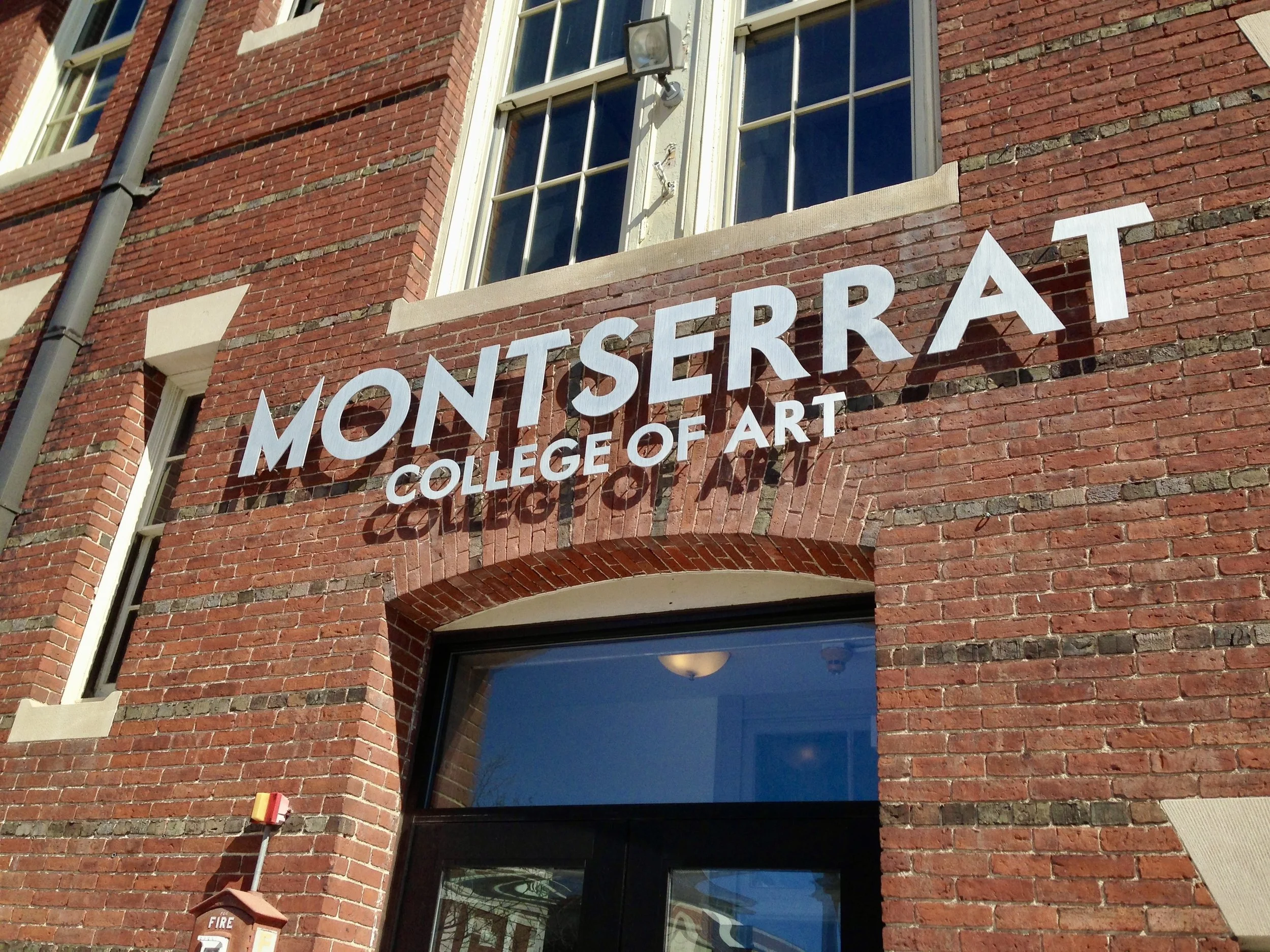Brick building entrance with signage reading “Montserr(at) College of Art,” windows, and glass doors.
