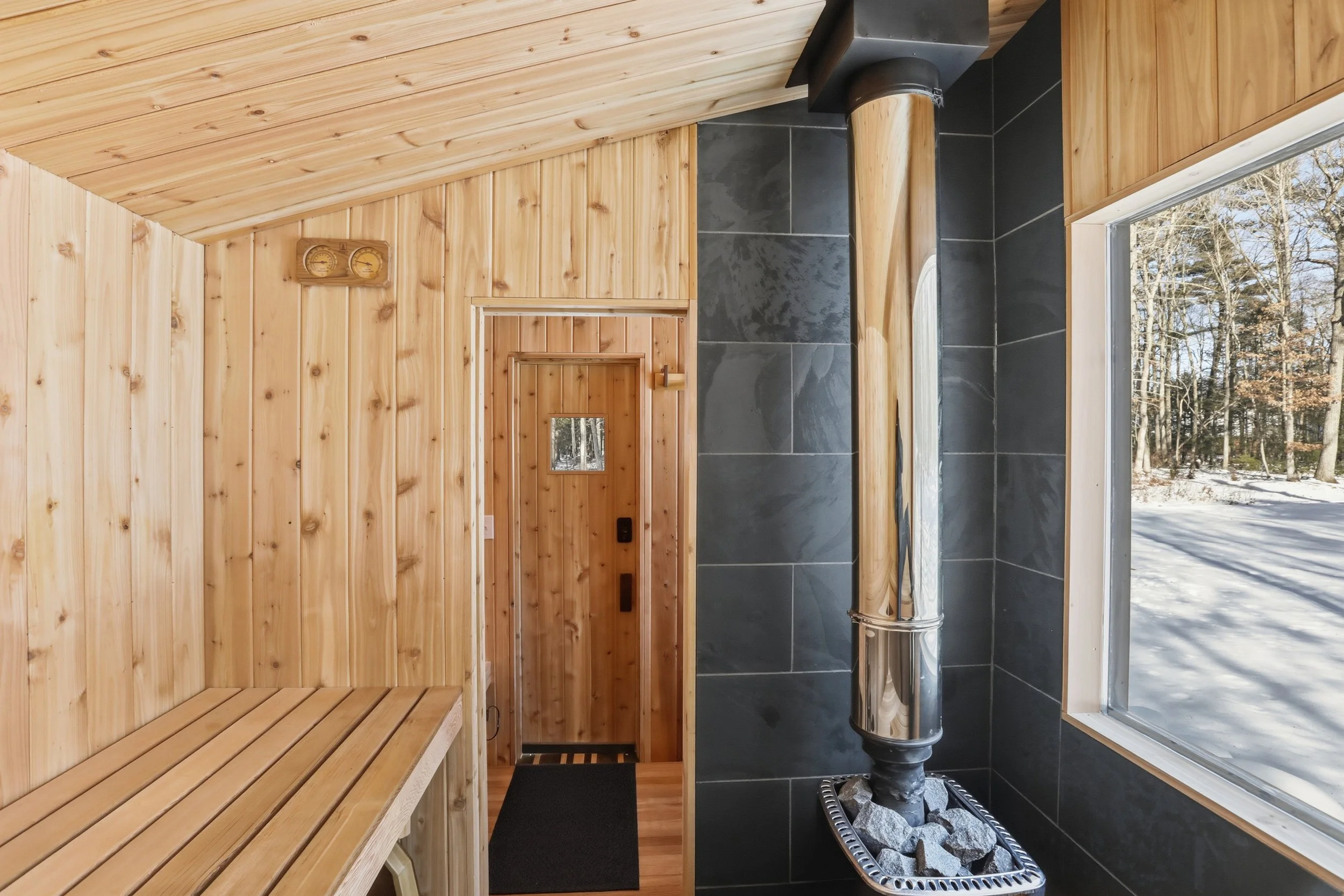 Wood-paneled sauna with benches, a black-tiled wood-burning stove, and a snowy forest visible through the window.