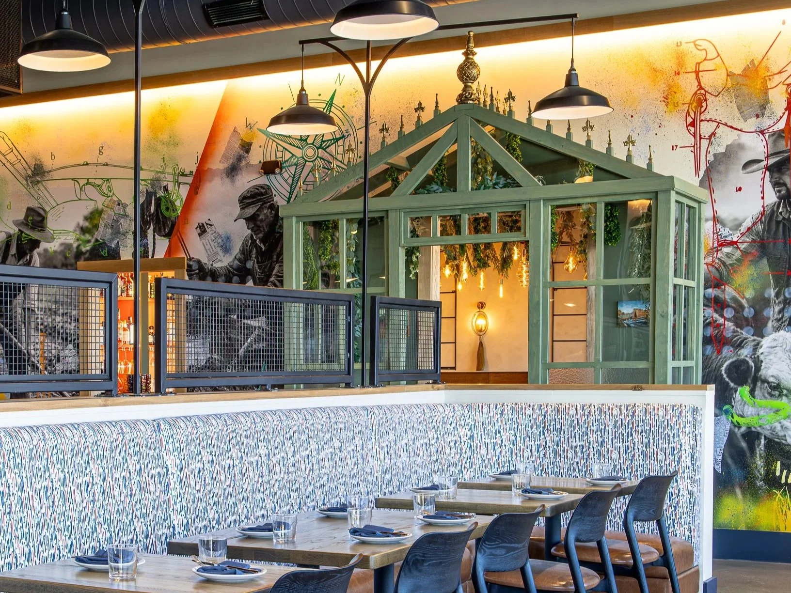 Modern restaurant interior with set dining tables, pendant lighting, colorful industrial artwork, and a green glass-enclosed dining area