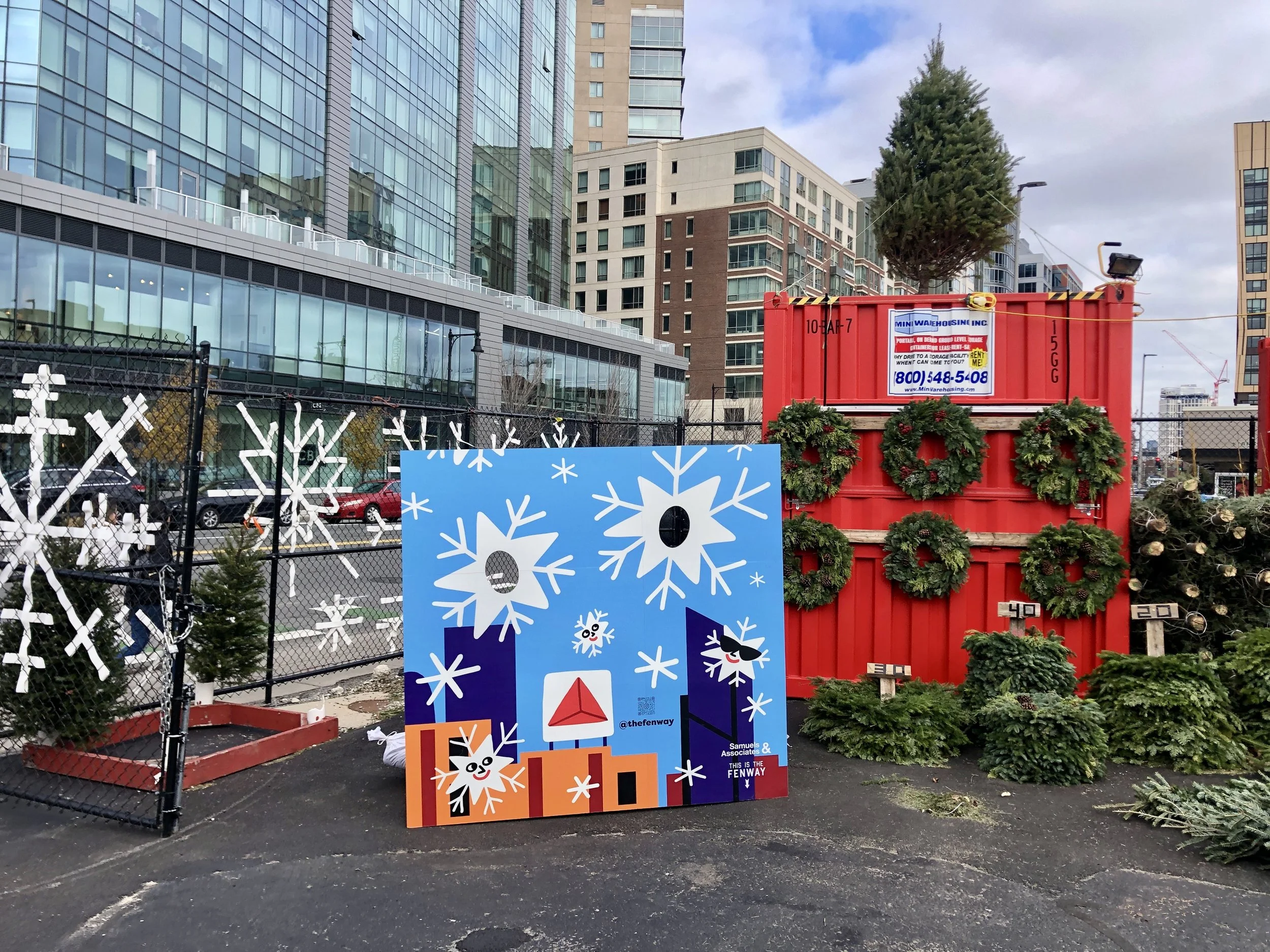 Holiday market display with Christmas wreaths, evergreen trees, snowflake decorations, and colorful photo board outside modern city buildings