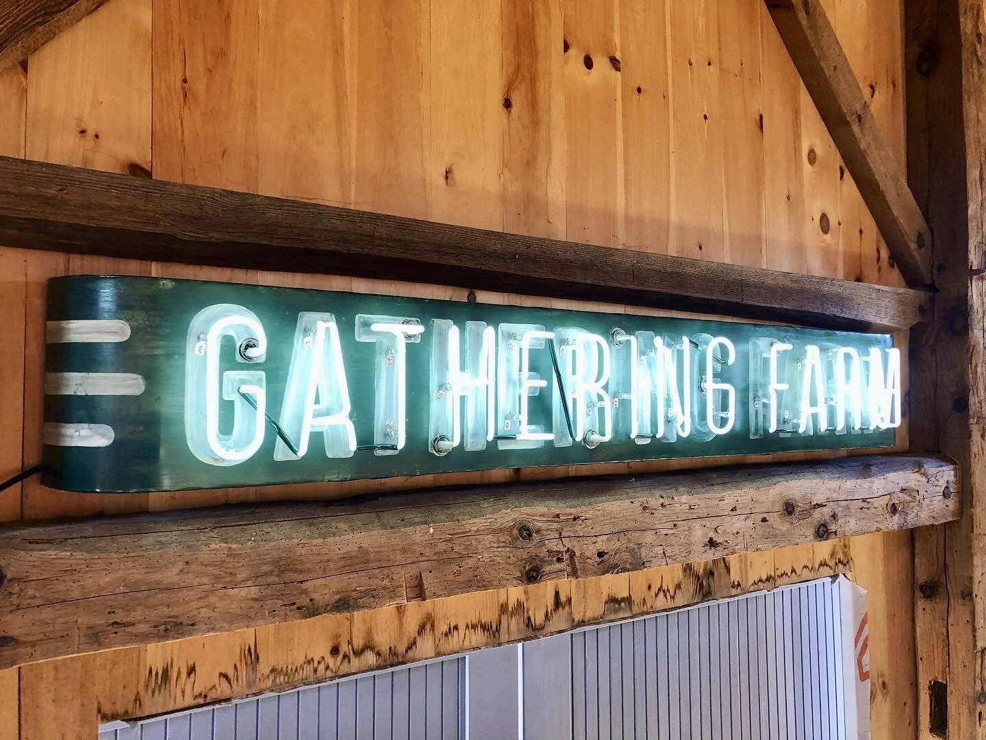 Illuminated neon sign reading “Gathering Farm” mounted on a rustic wooden barn wall