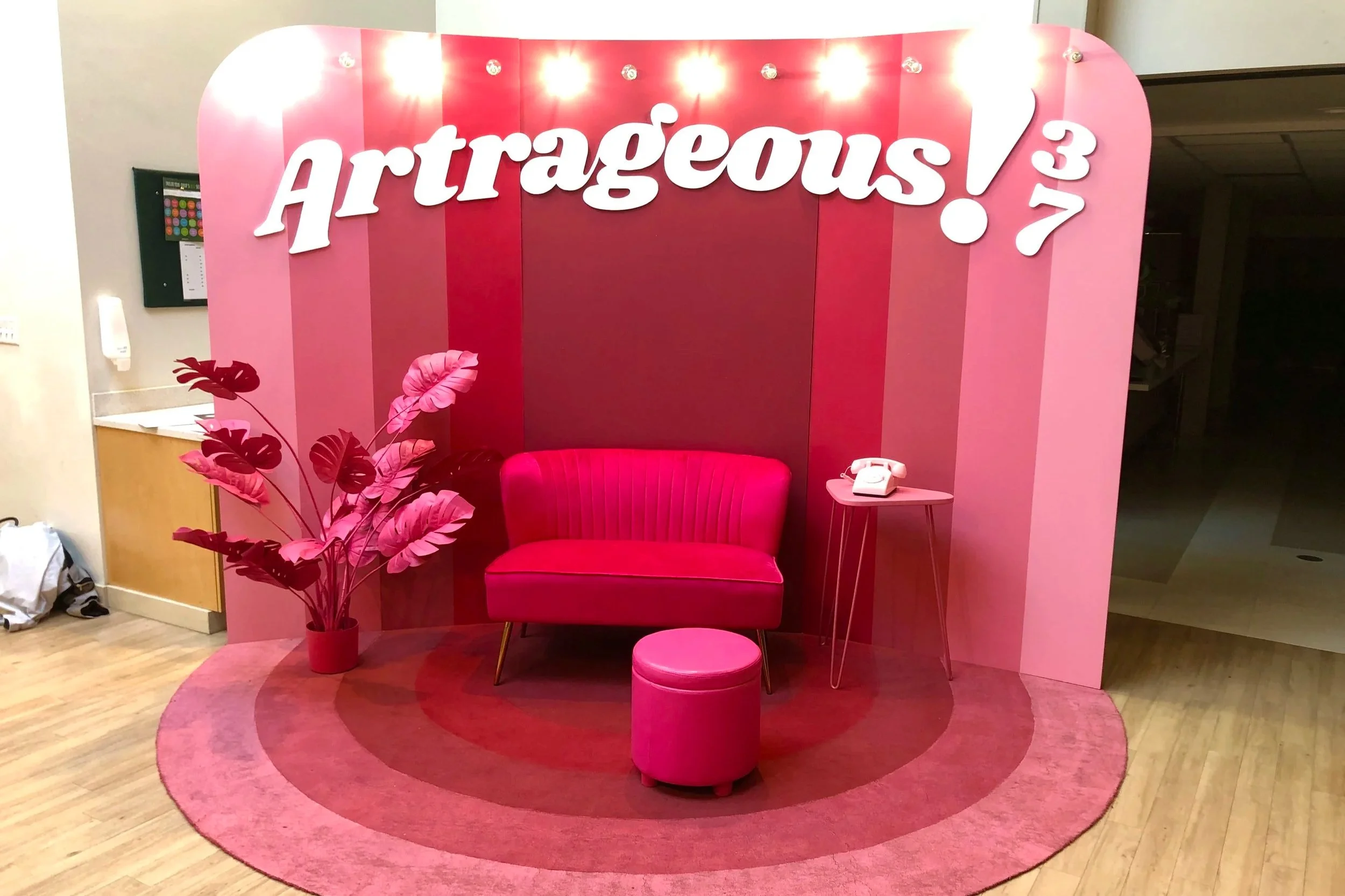 Pink retro-themed photo booth with a couch, ottoman, vintage telephone, potted tropical plant, striped backdrop, and illuminated “Atraeous” sign.