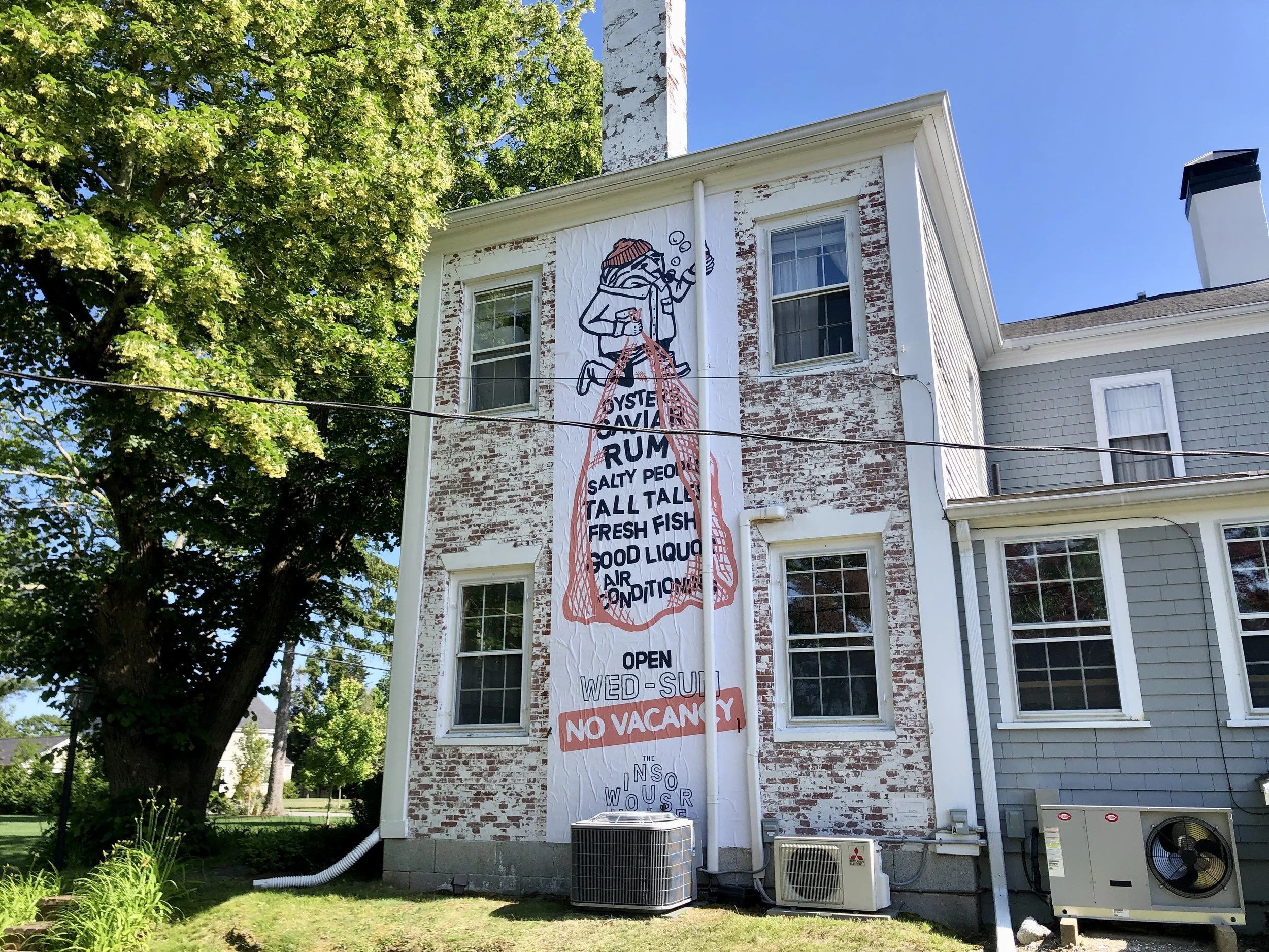 Historic brick building with peeling white paint, a large illustrated seafood restaurant banner, leafy tree, and outdoor air-conditioning units.
