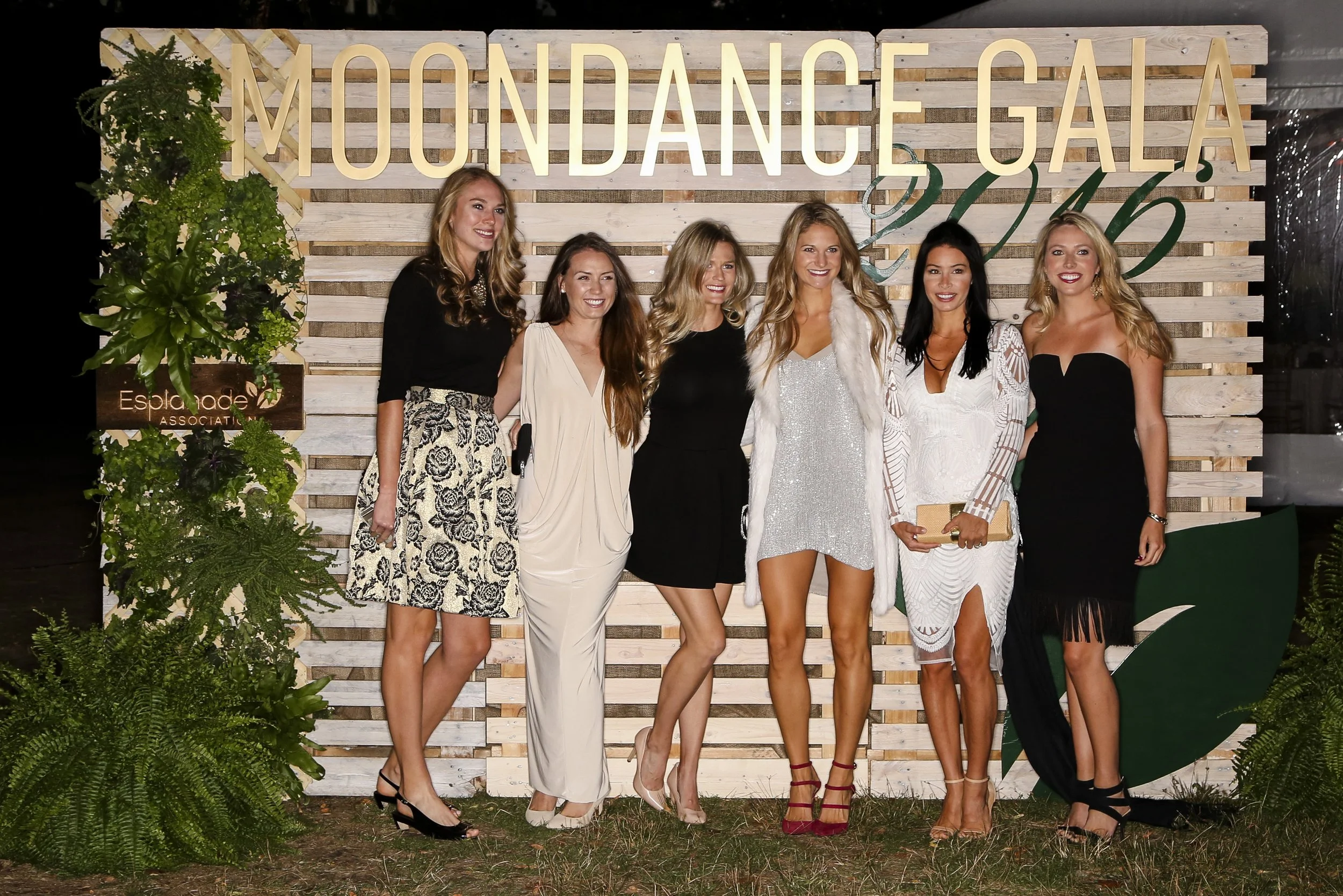 Six women posing in formal attire at a charity event in front of a wooden “MoonDance Gala” backdrop.