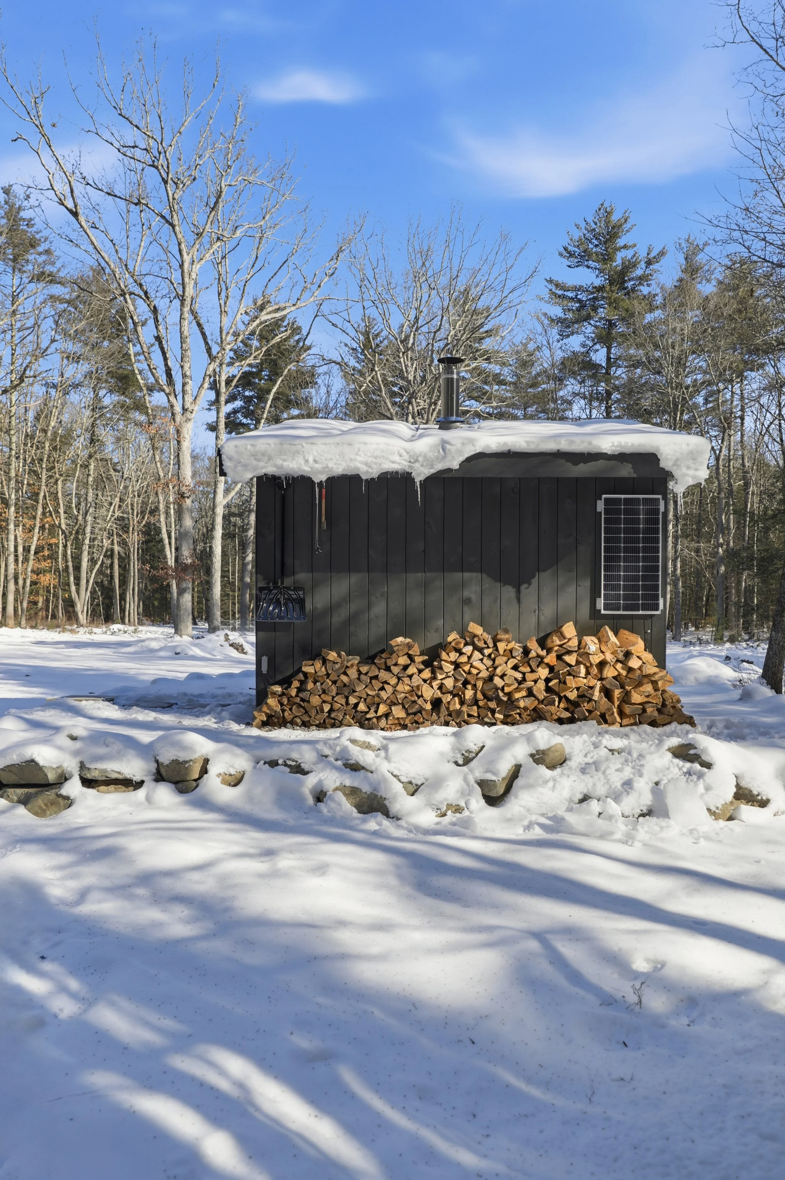 Black cabin with a snow-covered roof, chimney, solar panel, and stacked firewood in a snowy forest