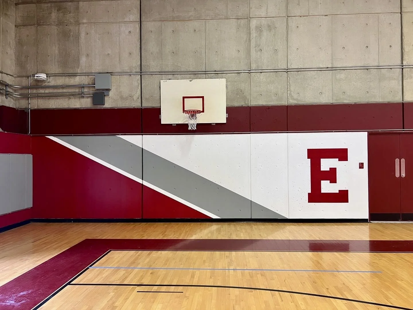 Indoor basketball court with a hoop, polished wood floor, and red-and-white wall featuring a large letter E