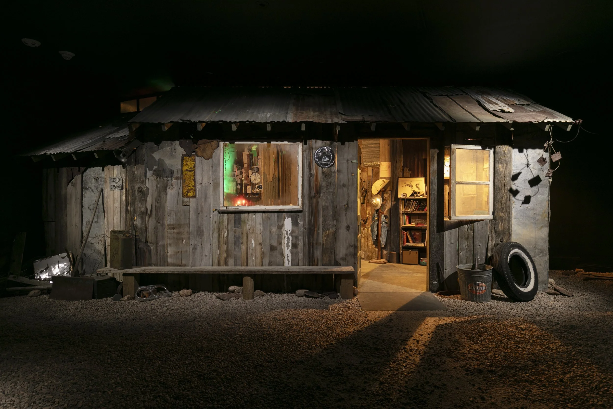 Rustic wooden shack at night with a corrugated metal roof, open doorway, lit window, bookshelves, and vintage outdoor objects