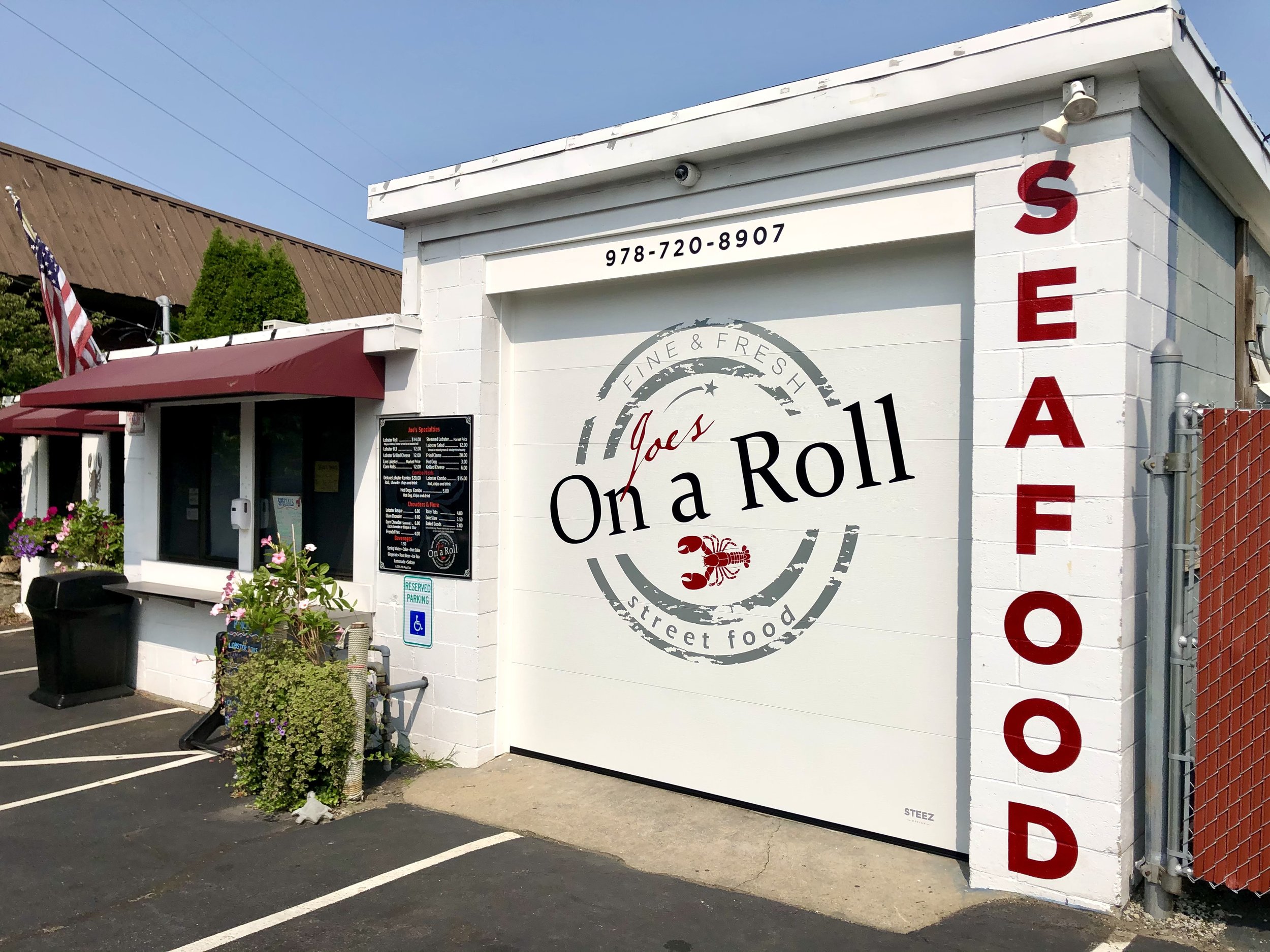 Exterior of Joe's On a Roll seafood restaurant with a lobster logo, menu board, American flag, and red awning.